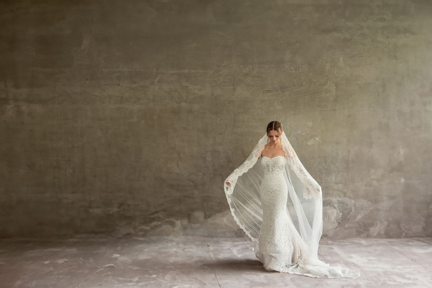 Ethereal bridal portrait with flowing veil against concrete wall.
