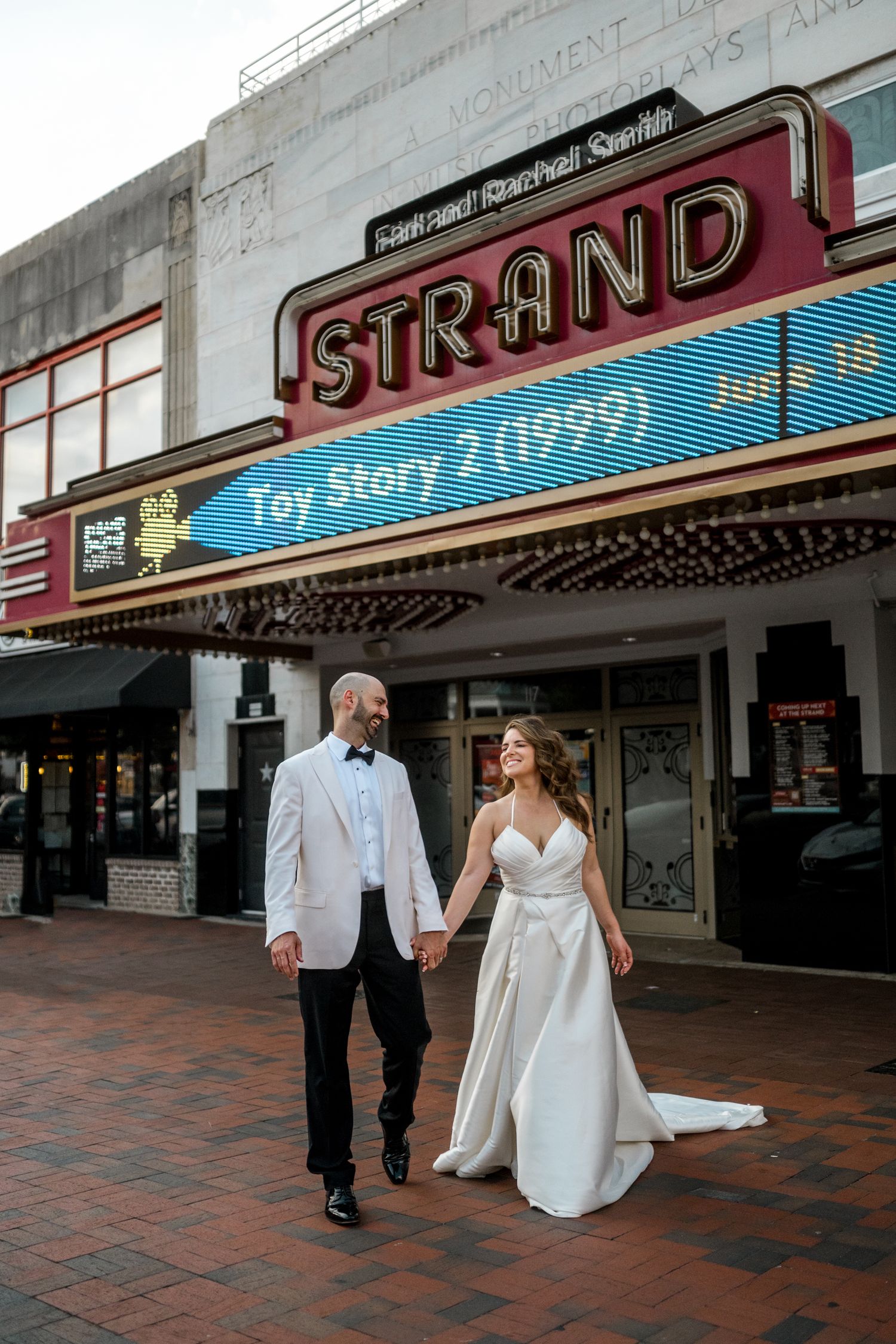 A couple in wedding attire walks together outside the historic Strand Theater with its vintage marquee sign visible.