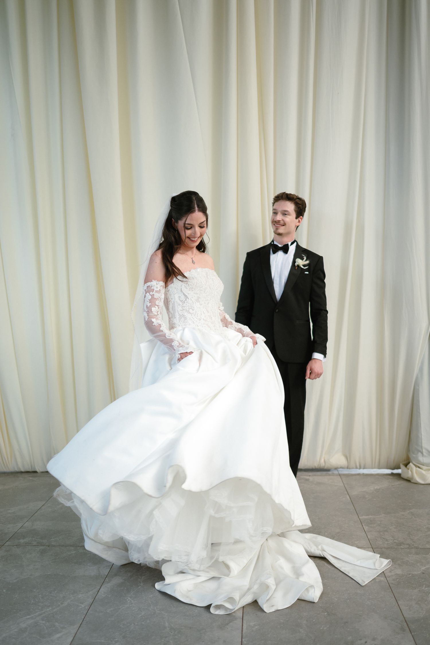Bride in white ballgown twirls while groom in black tuxedo smiles behind her against ivory drape backdrop.