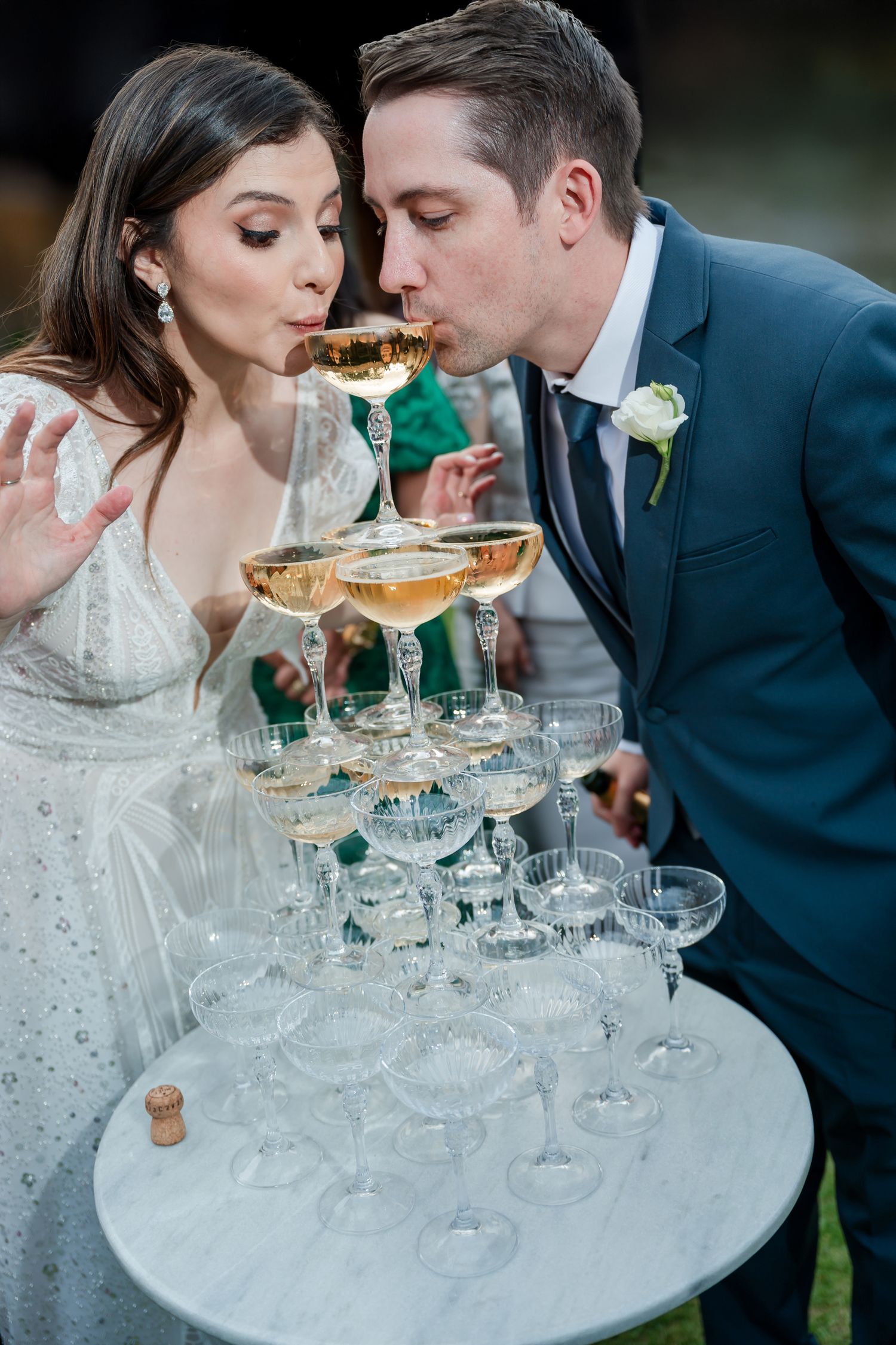 Newlyweds sipping champagne from a tower of stacked glasses outdoors.