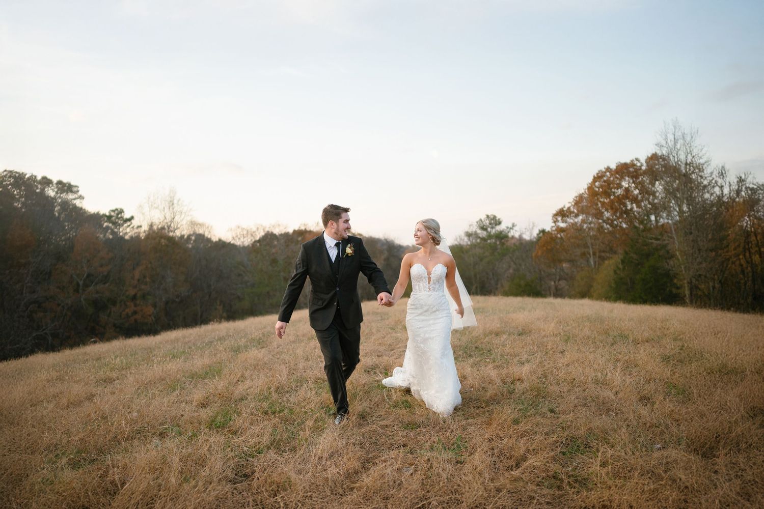 A couple in wedding attire walks together across a scenic hilltop during golden hour.