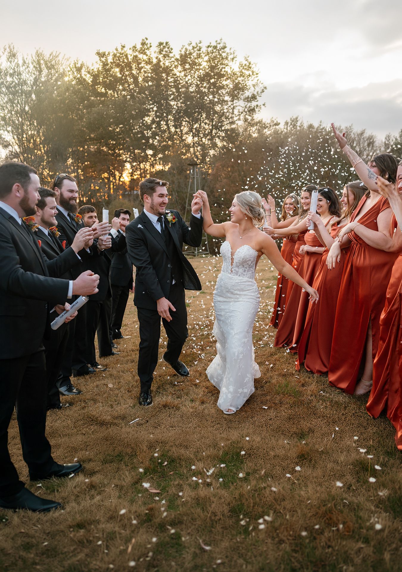 Bride and groom walk through confetti tunnel with wedding party in black suits and rust bridesmaid dresses at sunset.