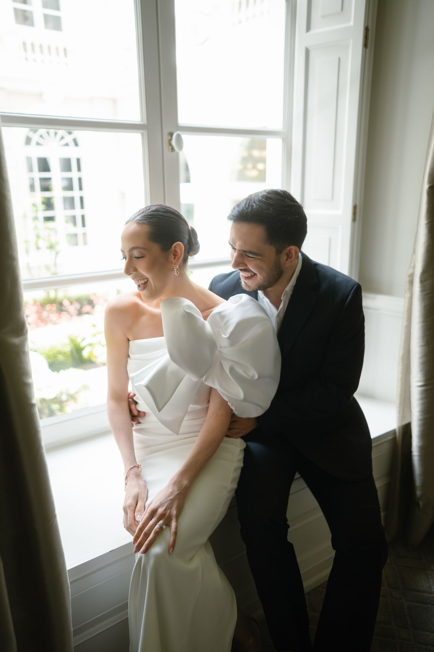 Joyful bride in white strapless gown embraced by groom in dark suit, laughing together by bright window.