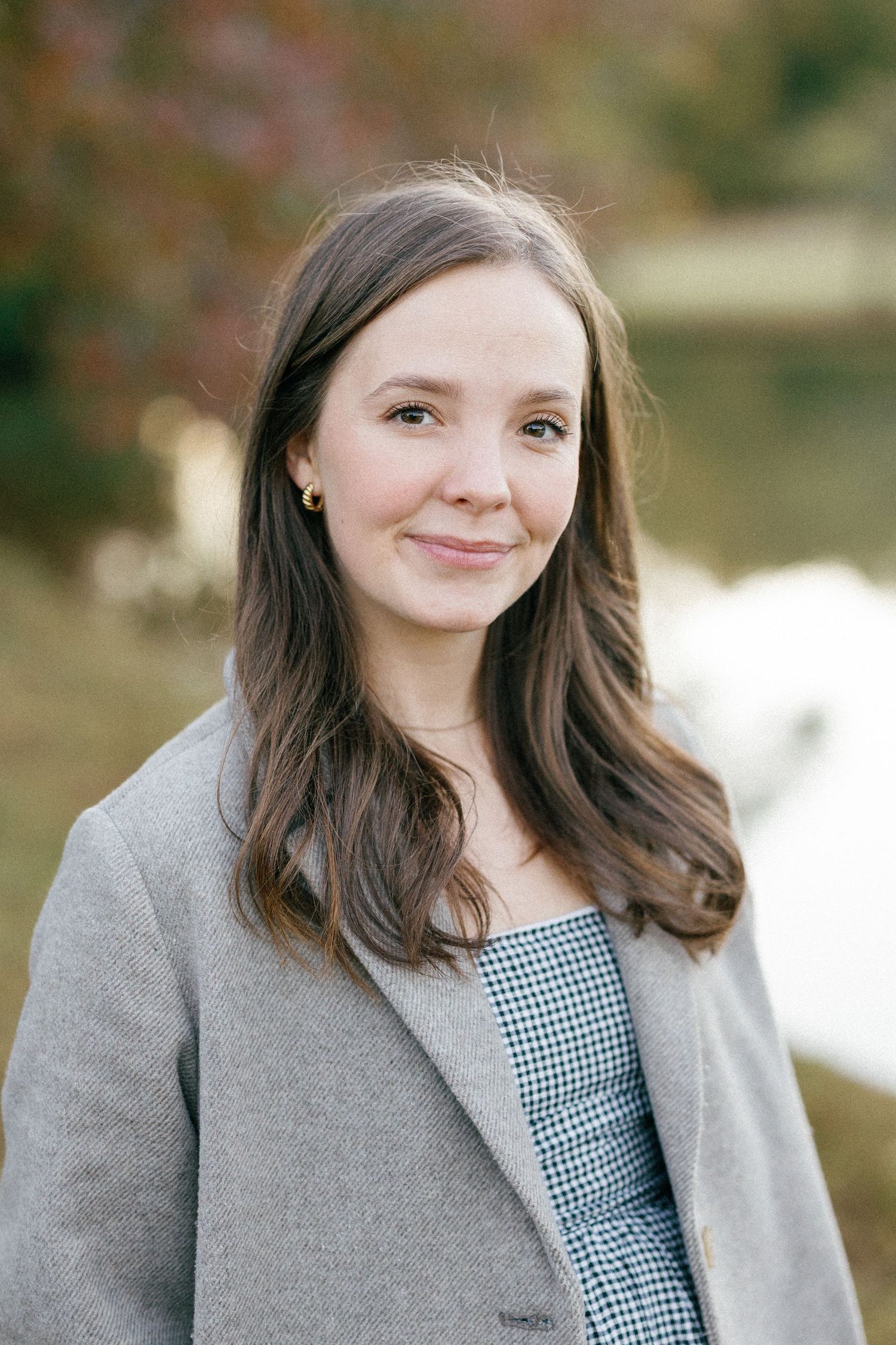 Portrait of someone wearing a gray cardigan over a gingham dress against a soft blurred outdoor background.