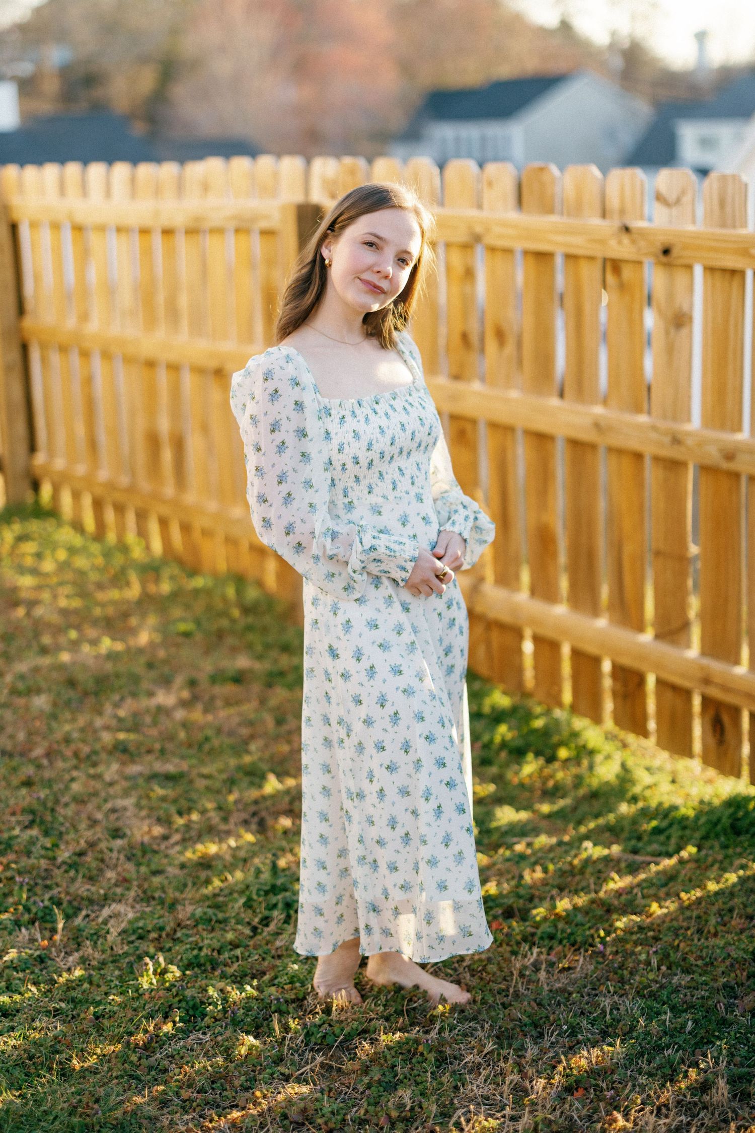 A person stands in a floral print maxi dress with long sleeves by a wooden fence on a sunny autumn day.