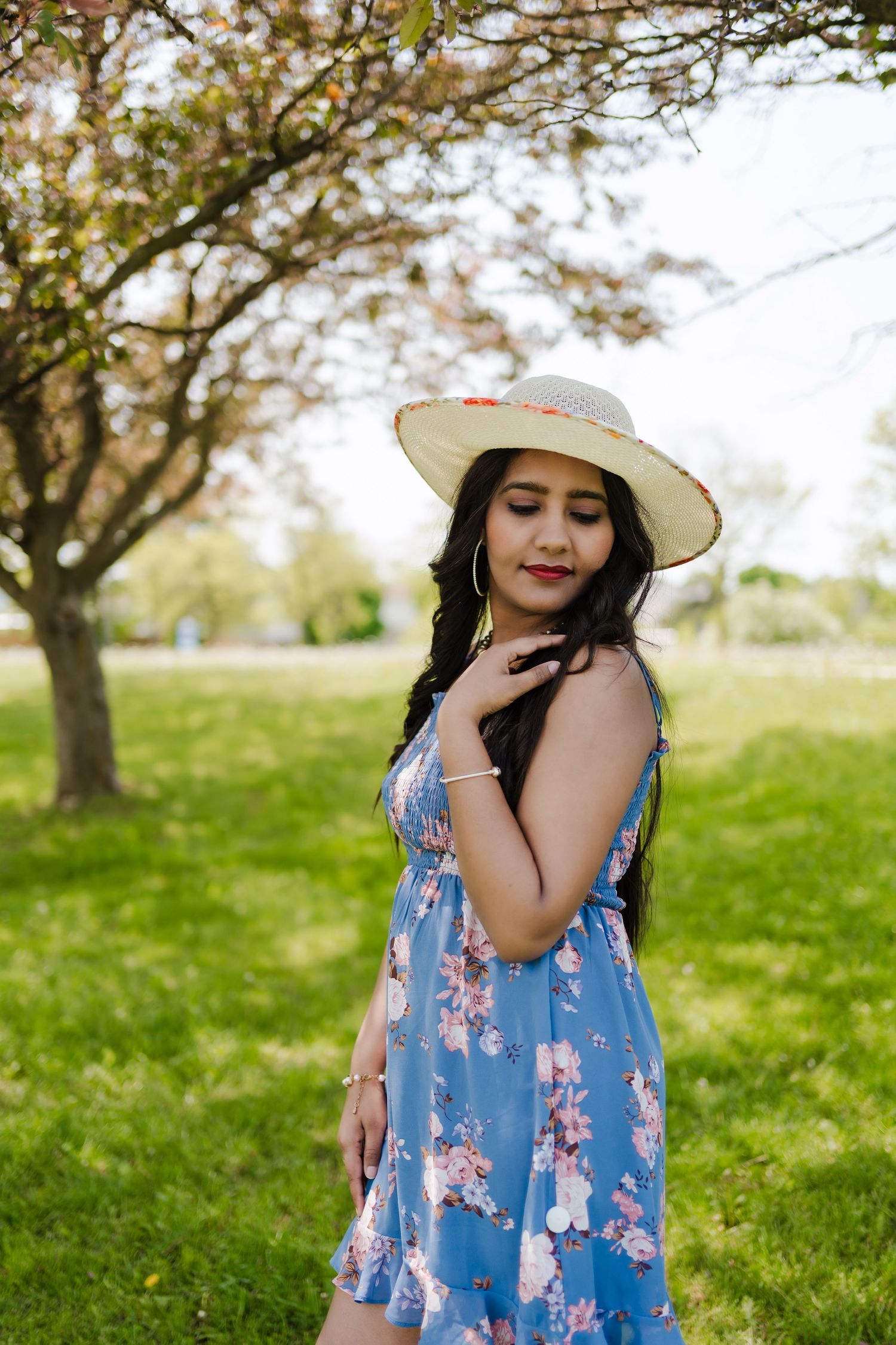Person in a blue floral dress and straw hat poses gracefully in a spring garden setting under blooming trees.