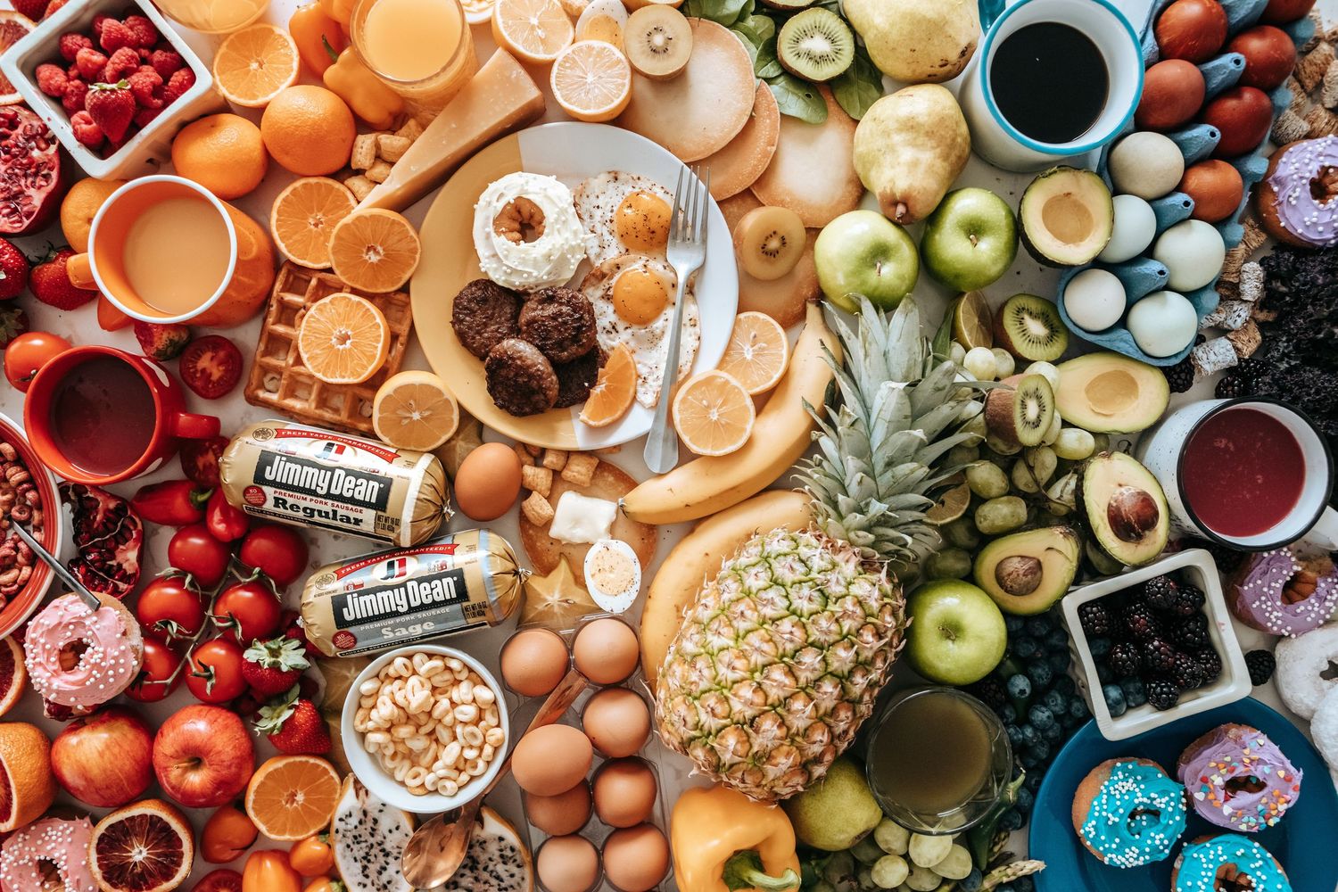 Colorful flat lay of assorted fresh fruits, vegetables, eggs, donuts, and foods arranged in a rainbow pattern.
