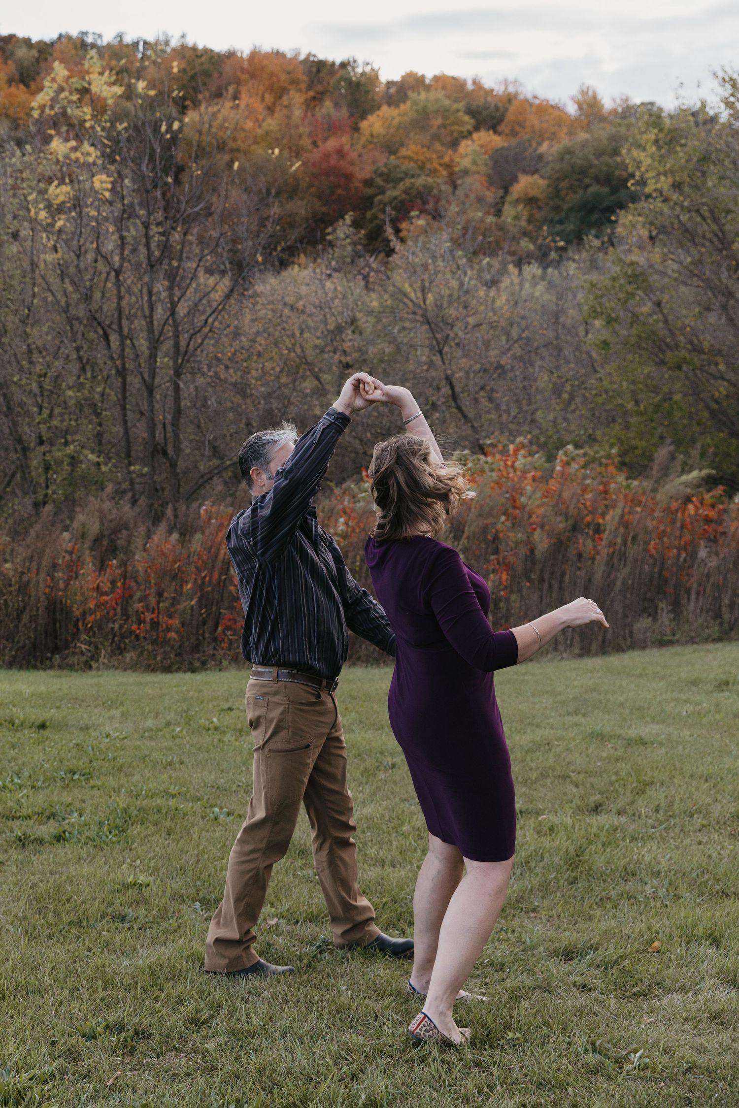 Couple dancing outdoors in autumn field with colorful fall foliage trees in background.