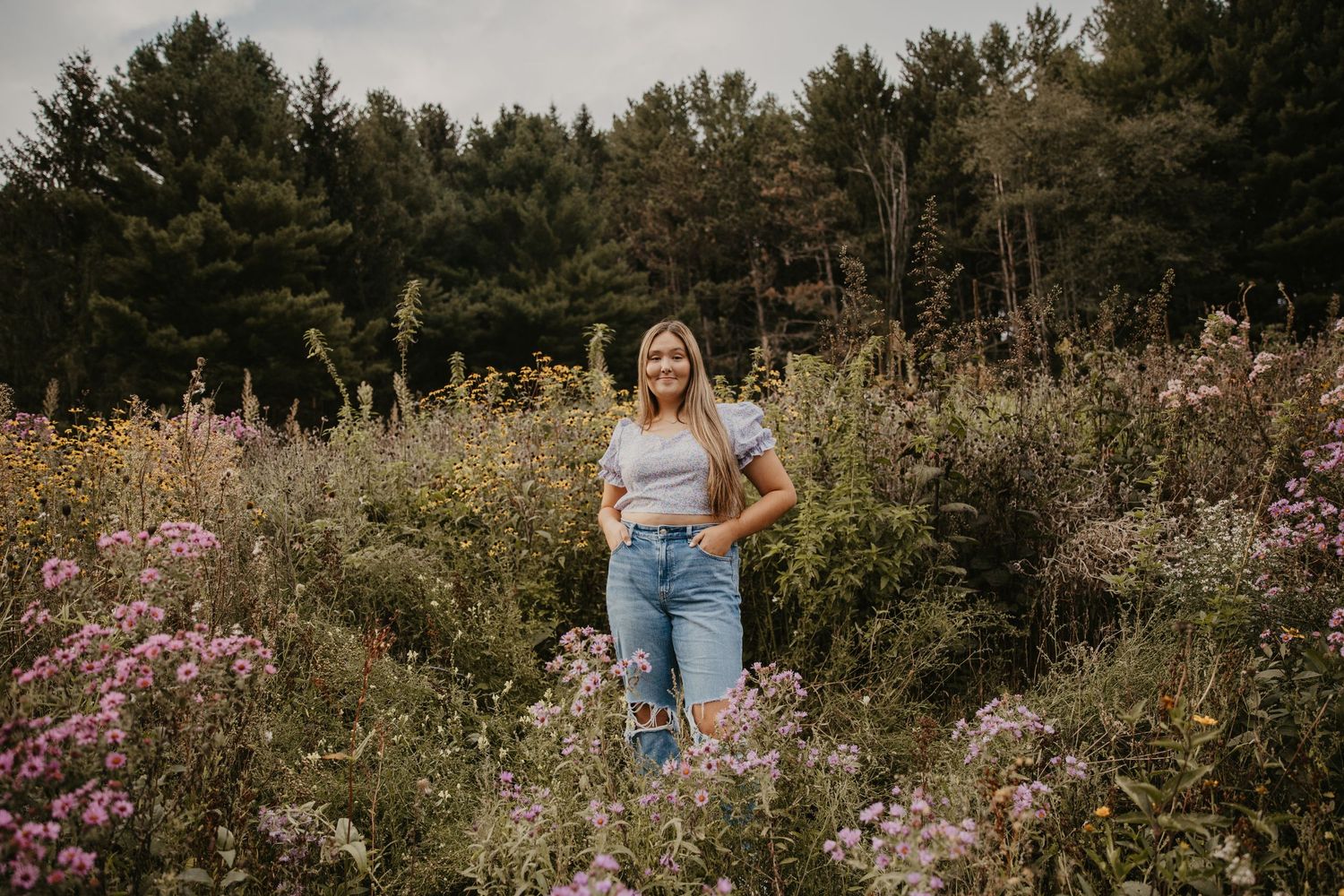Young woman in jeans and gray top standing among wildflowers in a lush meadow with evergreen trees behind her.