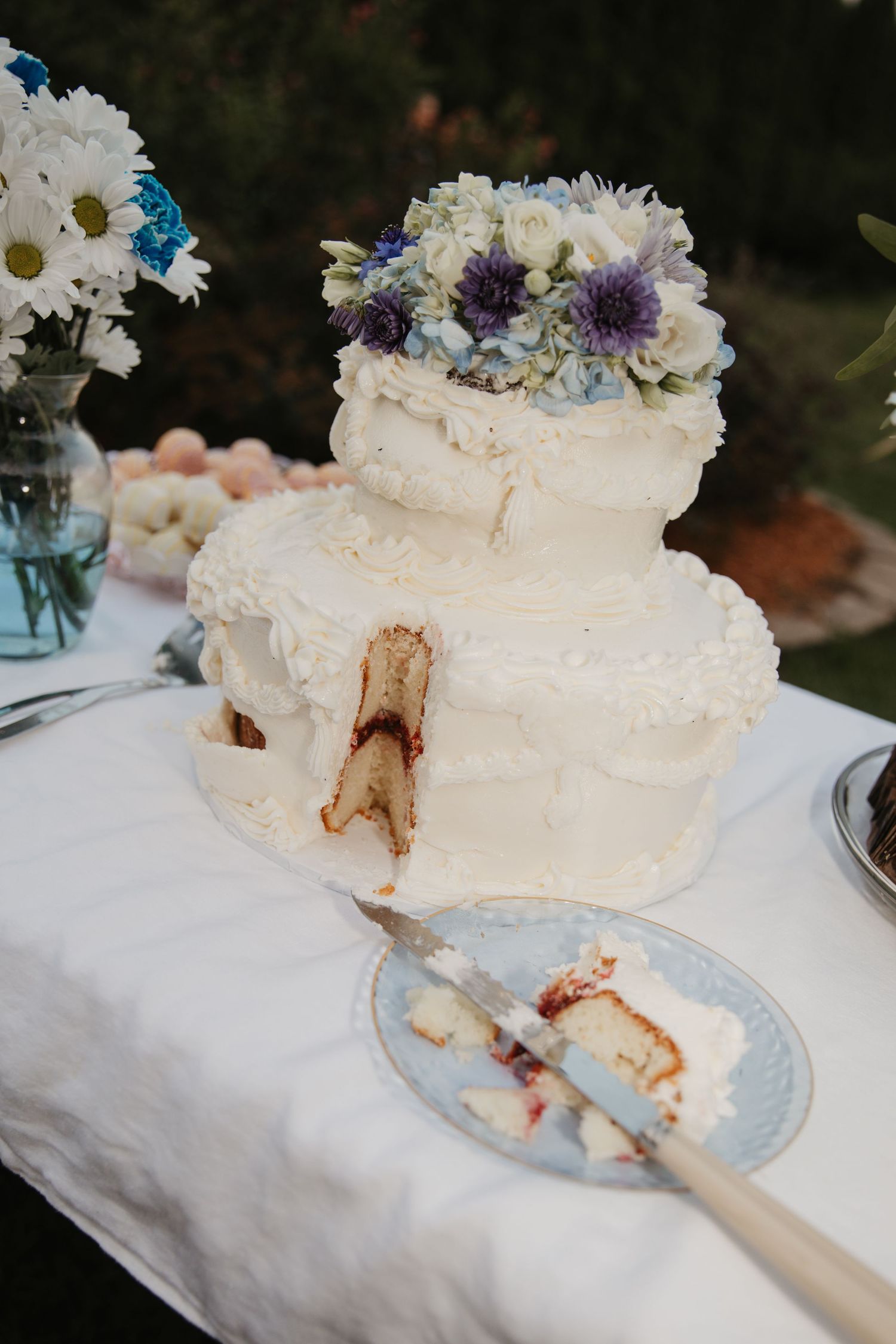 Two-tier white frosted wedding cake topped with blue and white flowers, with a slice cut out revealing red jam filling.