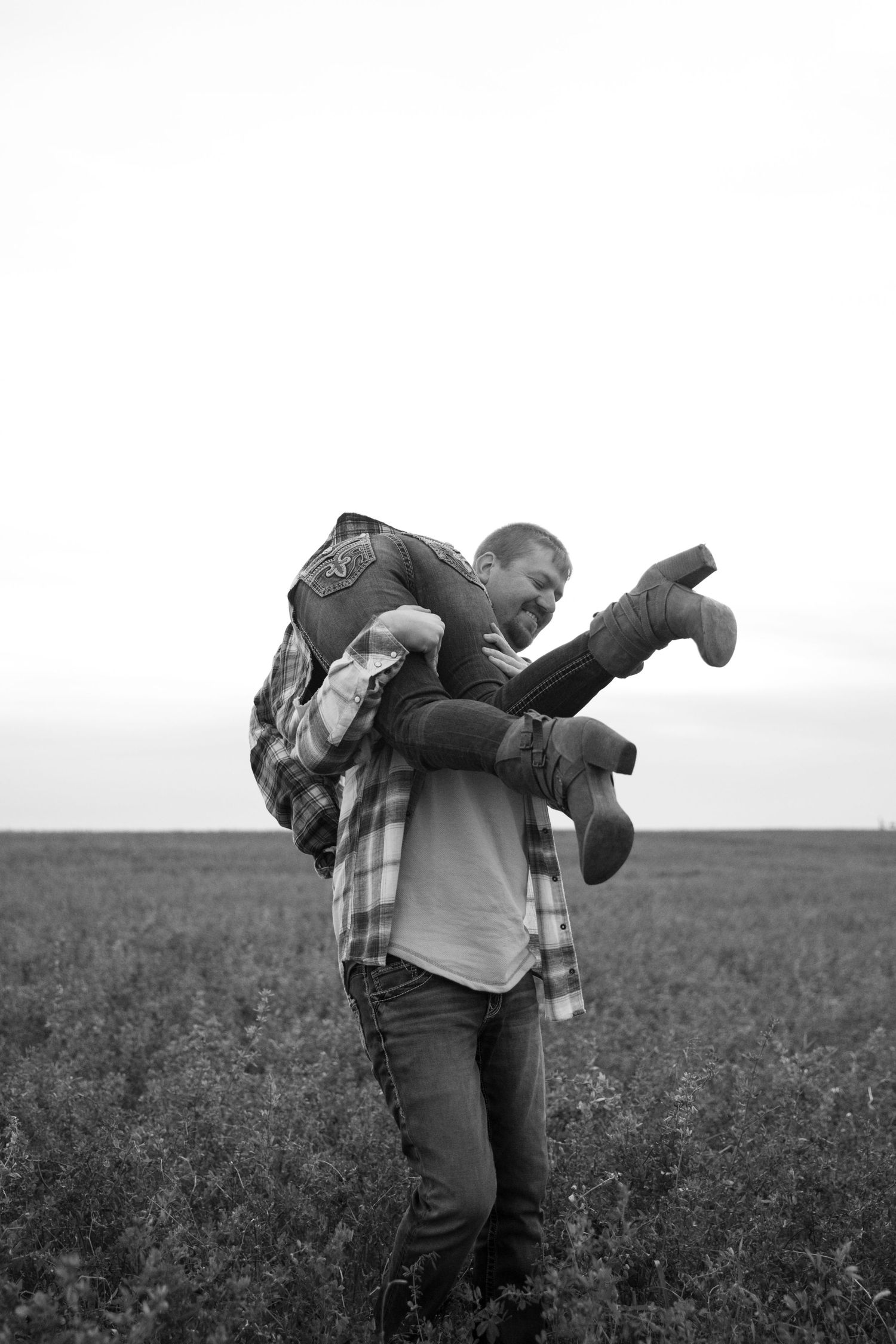 Black and white photo of a man carrying a child on his back in an open field, sharing a joyful outdoor moment.