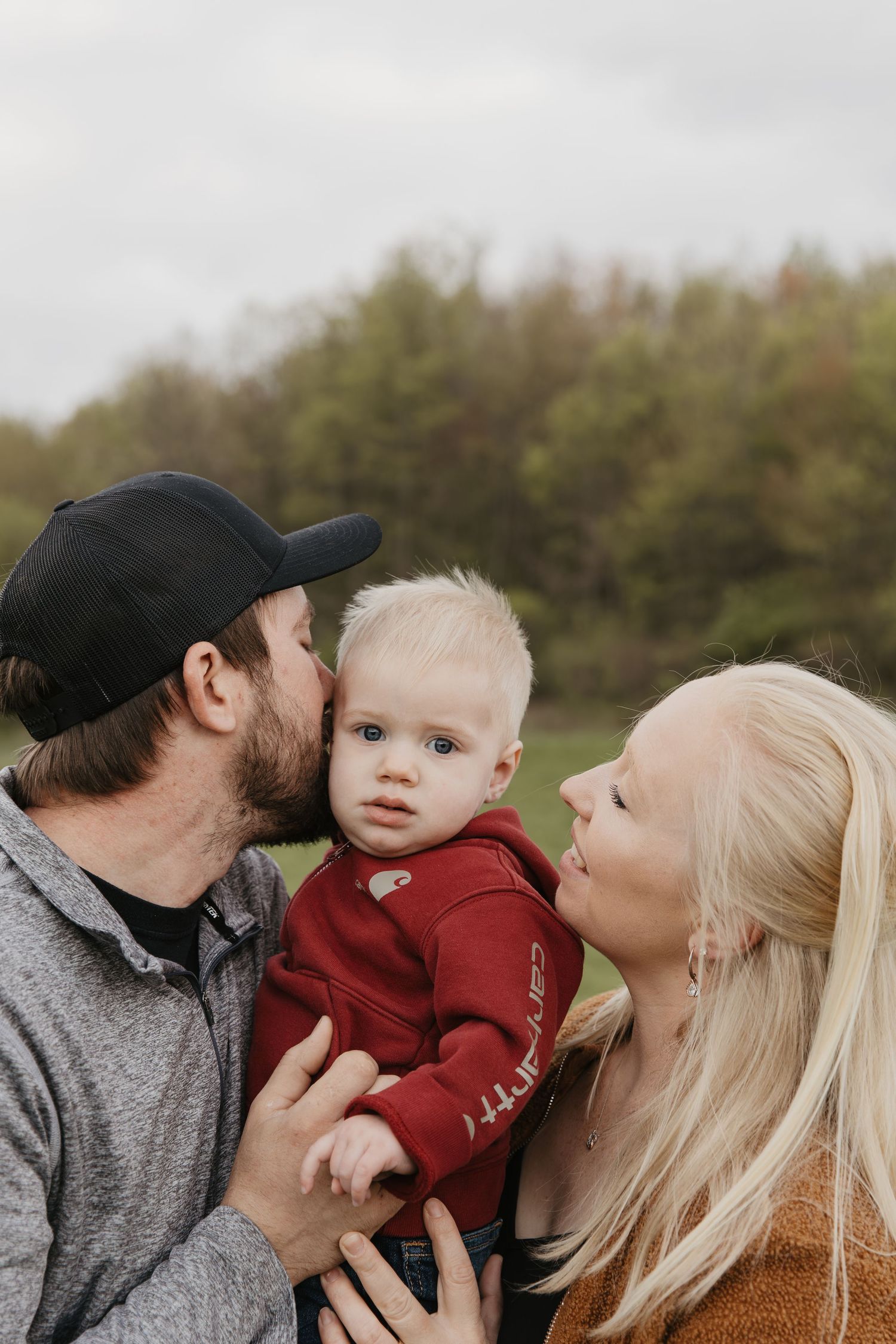 A young couple kisses their blonde toddler boy in a red hoodie outdoors with green trees in the background.