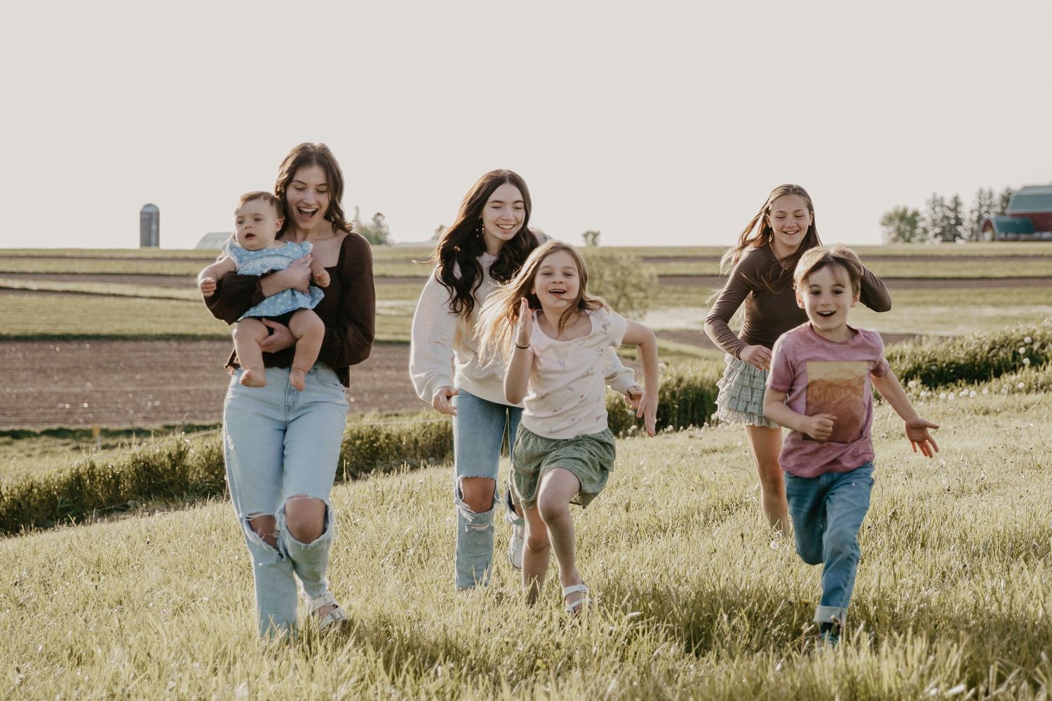 Women and children running joyfully through a green field on a sunny day in a rural setting.