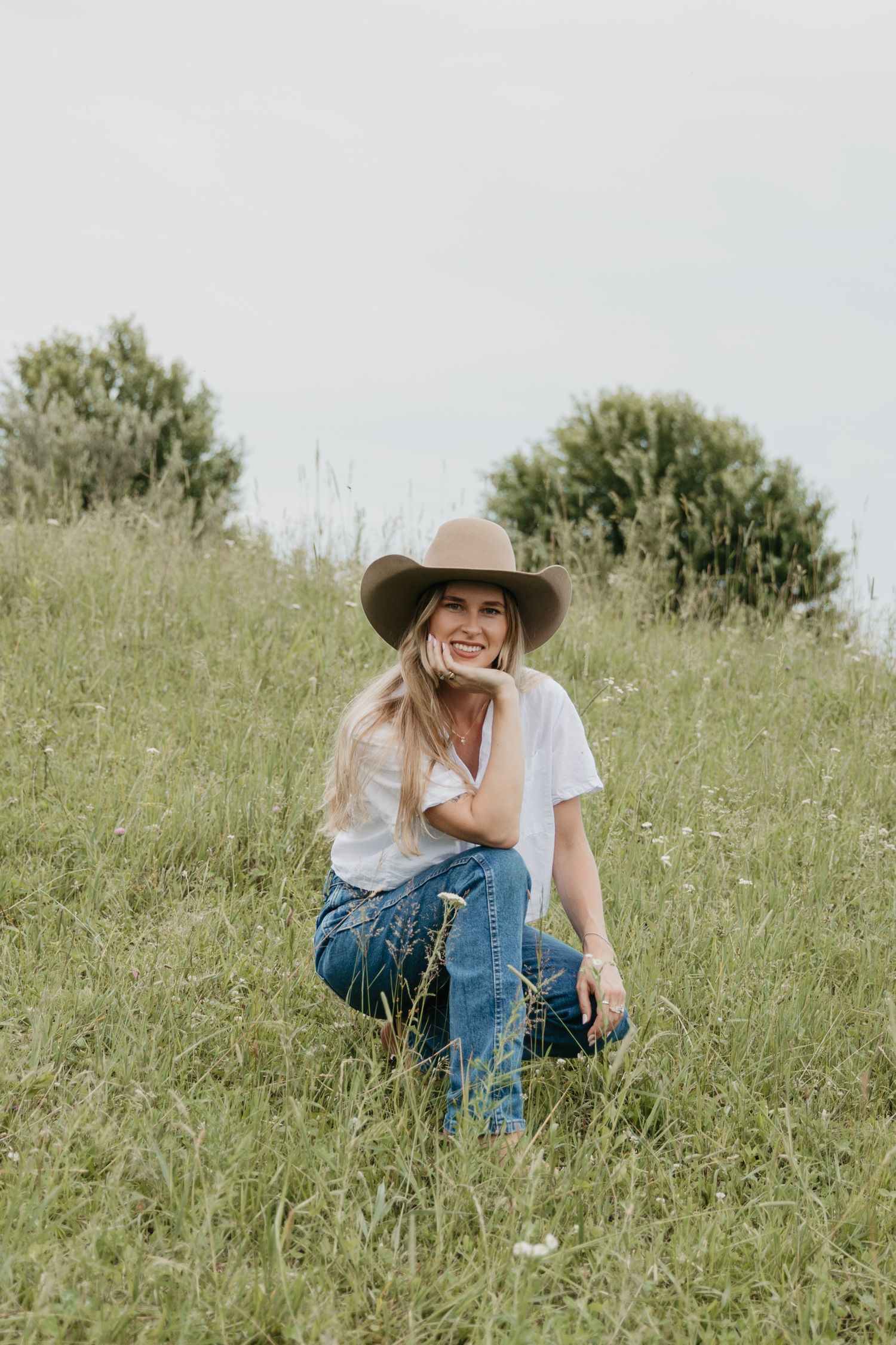Smiling blonde woman in white tee, jeans, and tan cowboy hat sitting in a lush green grassy field.