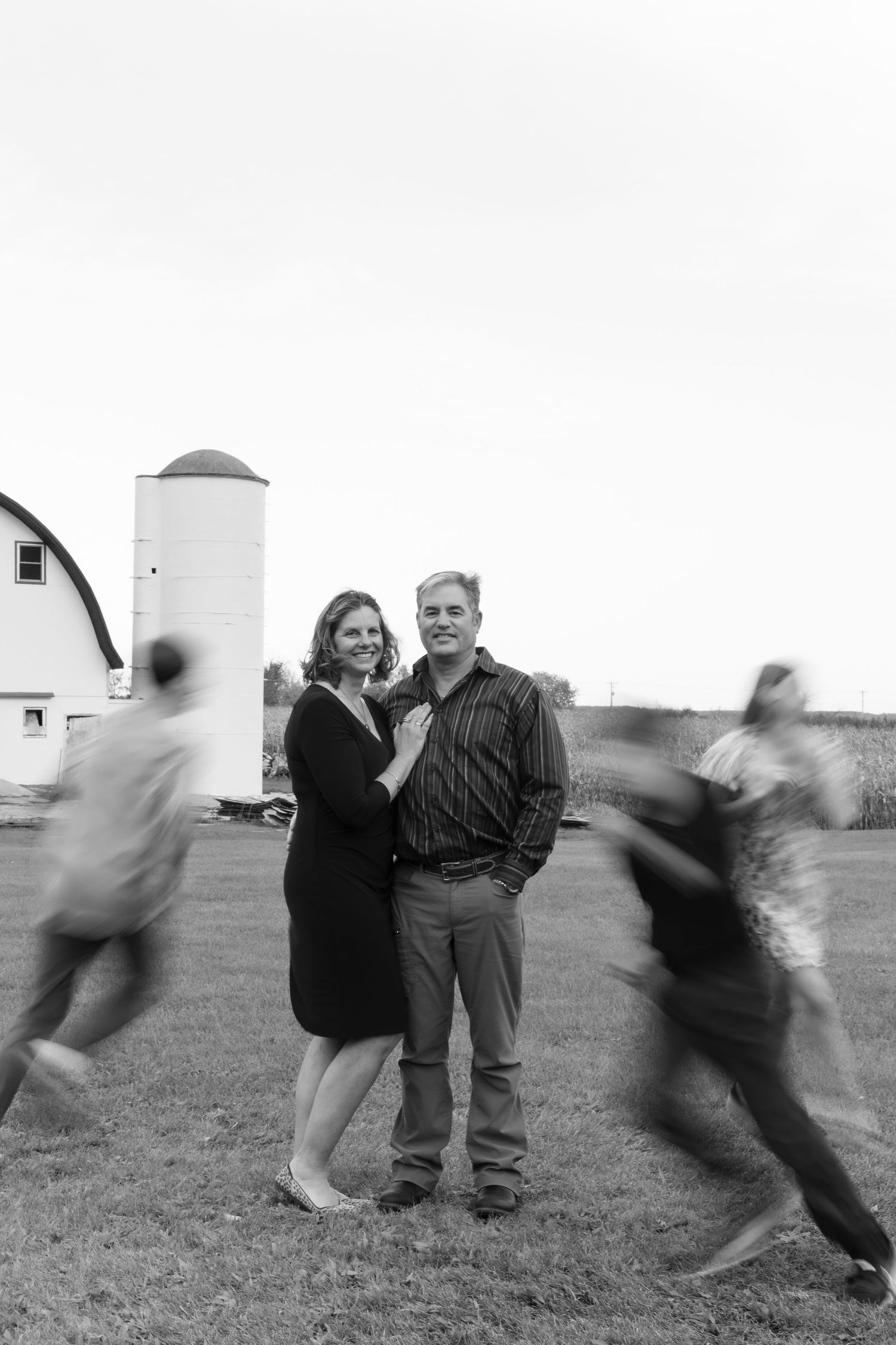 Couple posing by white barn and silo with blurred figures in black and white photo.