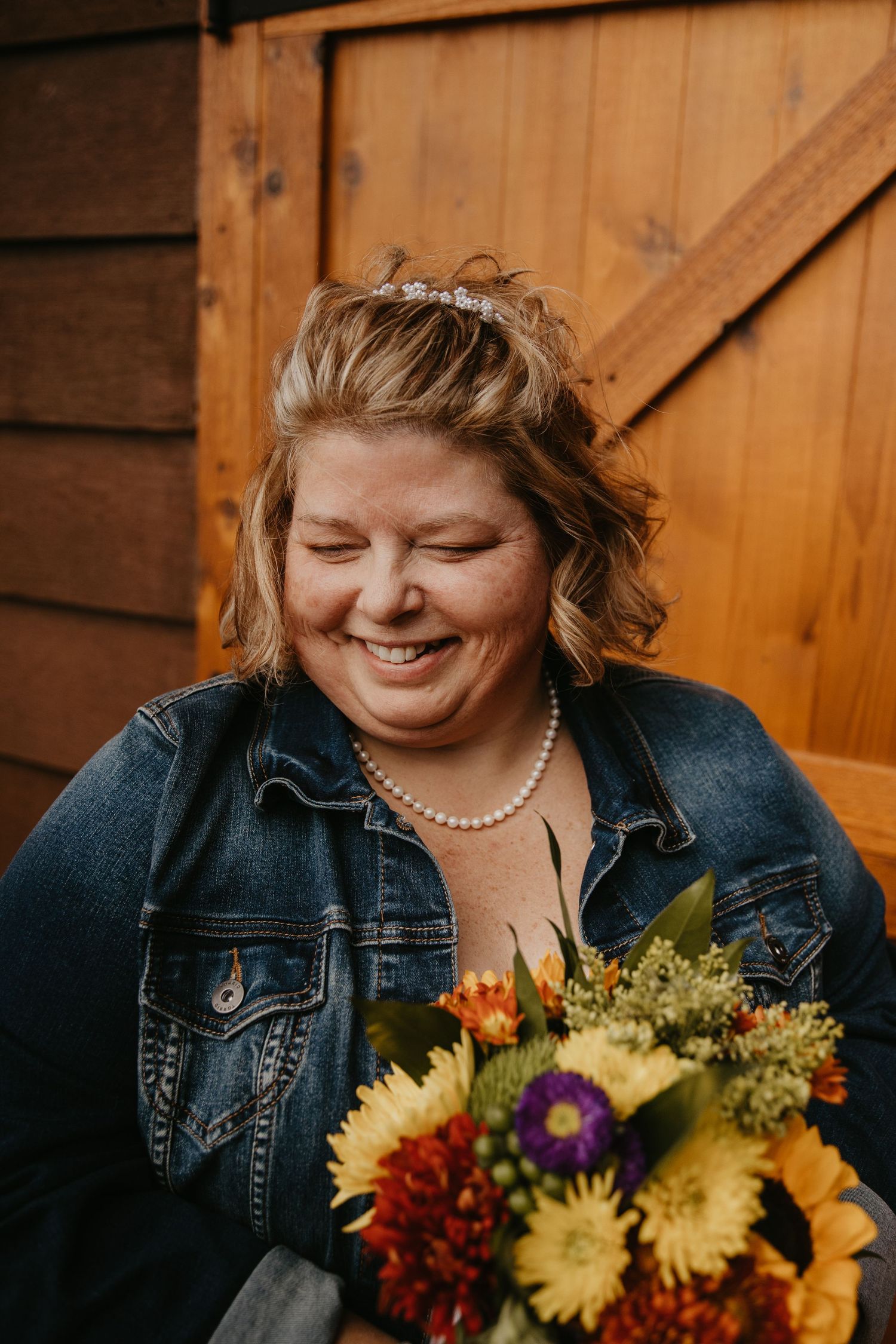 Smiling bride in denim jacket and pearl necklace holds colorful fall bouquet with sunflowers before wooden barn backdrop.