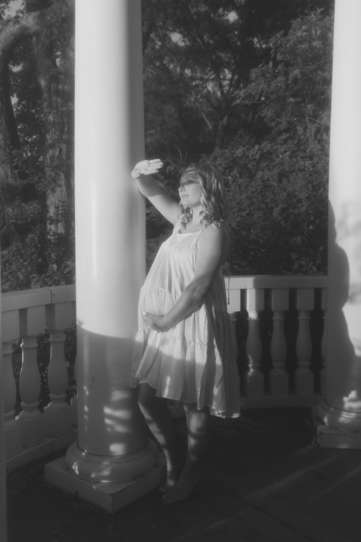 Black and white photo of a pregnant woman in a white dress standing on a porch, cradling her belly.