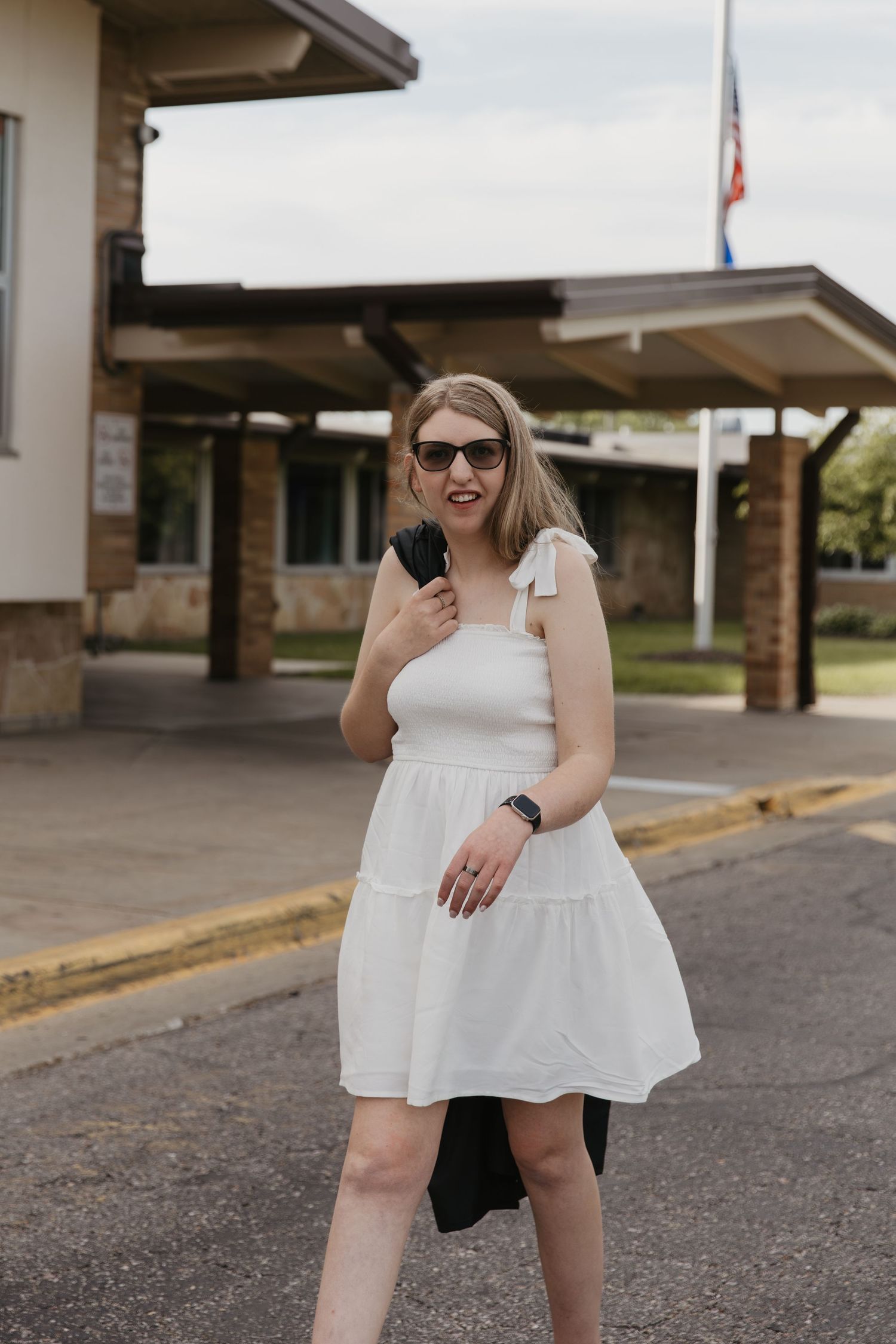 Woman in white sundress and sunglasses posing outdoors near a building with an American flag in background.