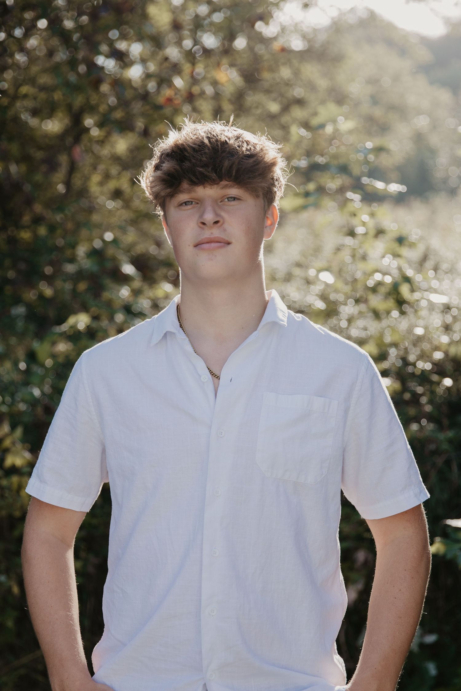 A young man in a white short-sleeve button-up shirt poses outdoors with green trees in the background.