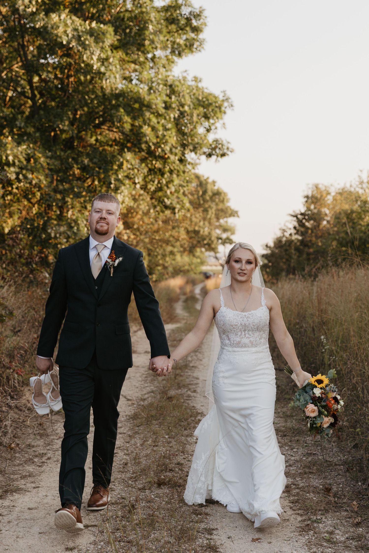 Bride and groom walk hand-in-hand down a rustic dirt path surrounded by autumn trees, she holds a sunflower bouquet.