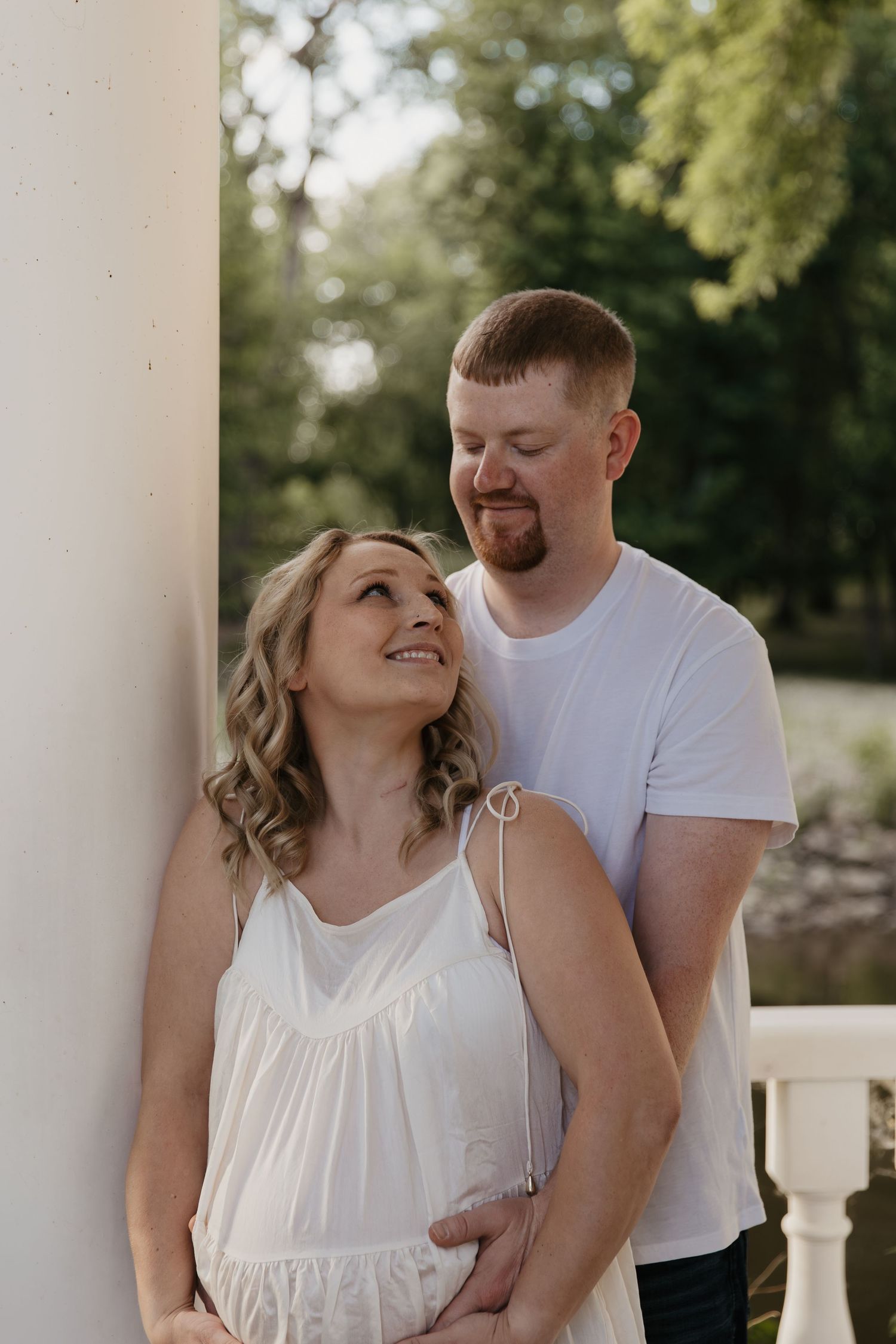 A smiling couple shares a tender moment on a sunlit porch, the woman in white dress gazing up at her bearded partner.