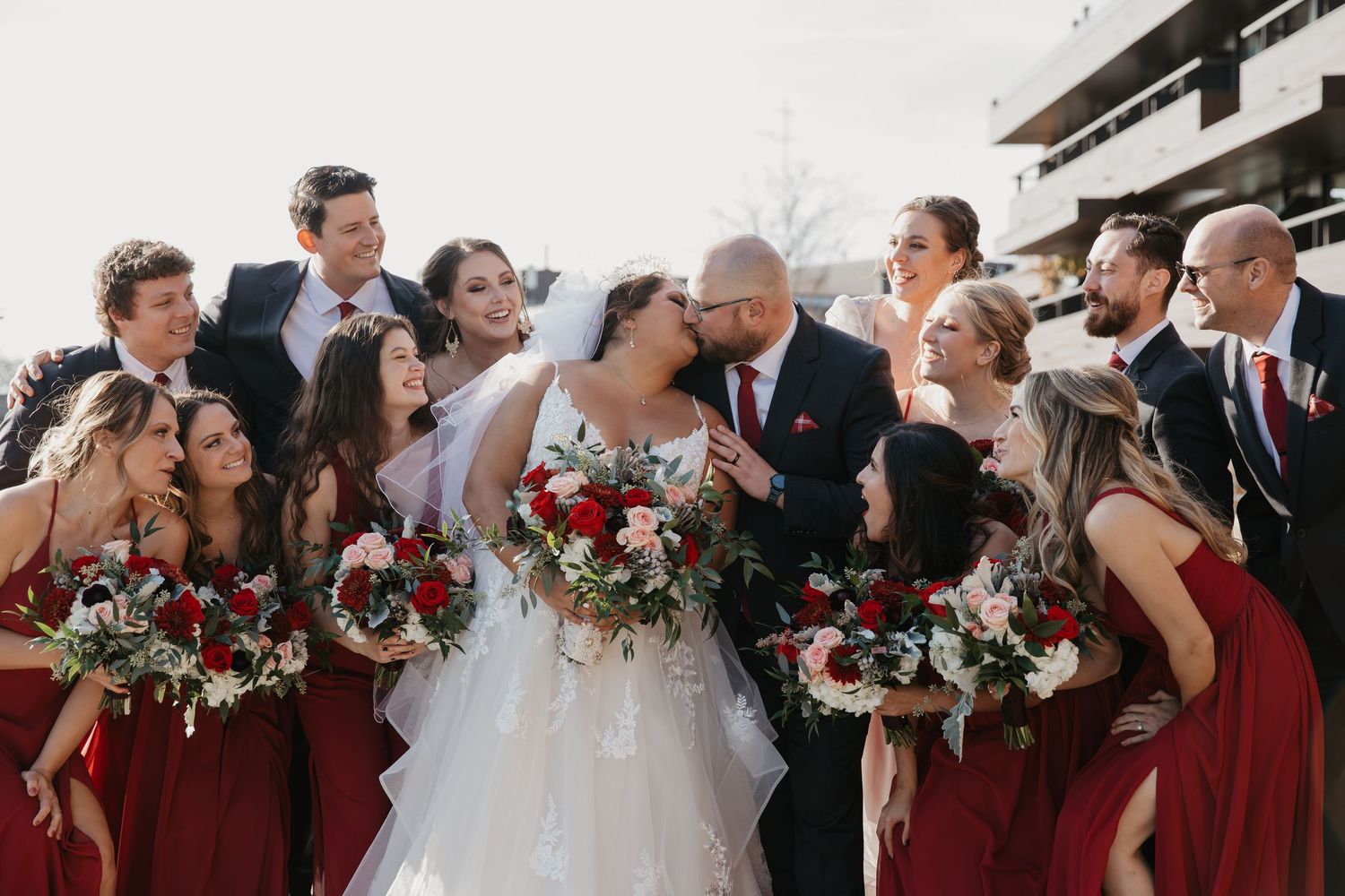 Bride and groom kiss surrounded by wedding party in burgundy dresses and dark suits holding red floral bouquets outdoors.