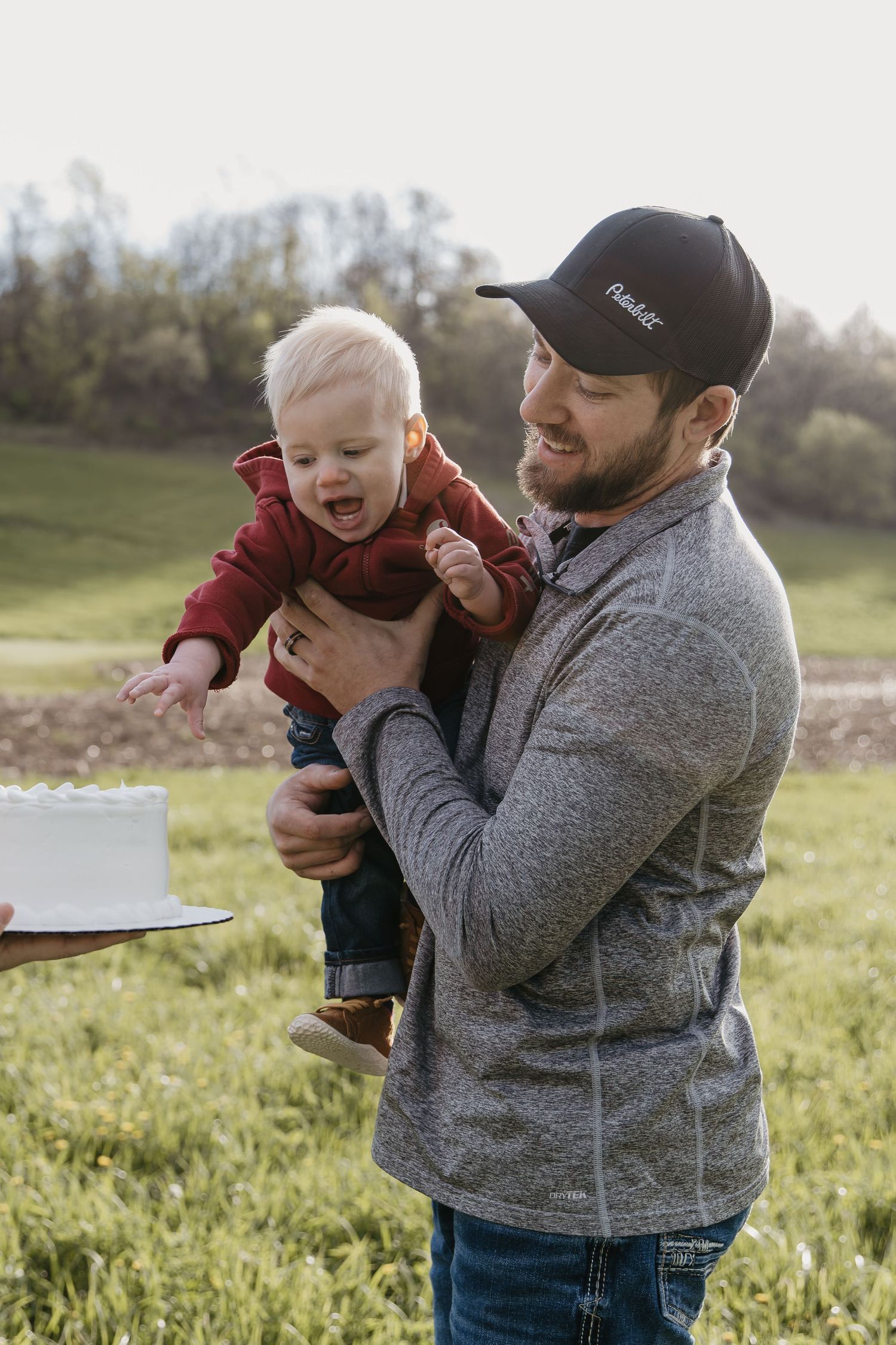 A father holds his baby near a white cake outdoors during a first birthday smash cake celebration in a green field.