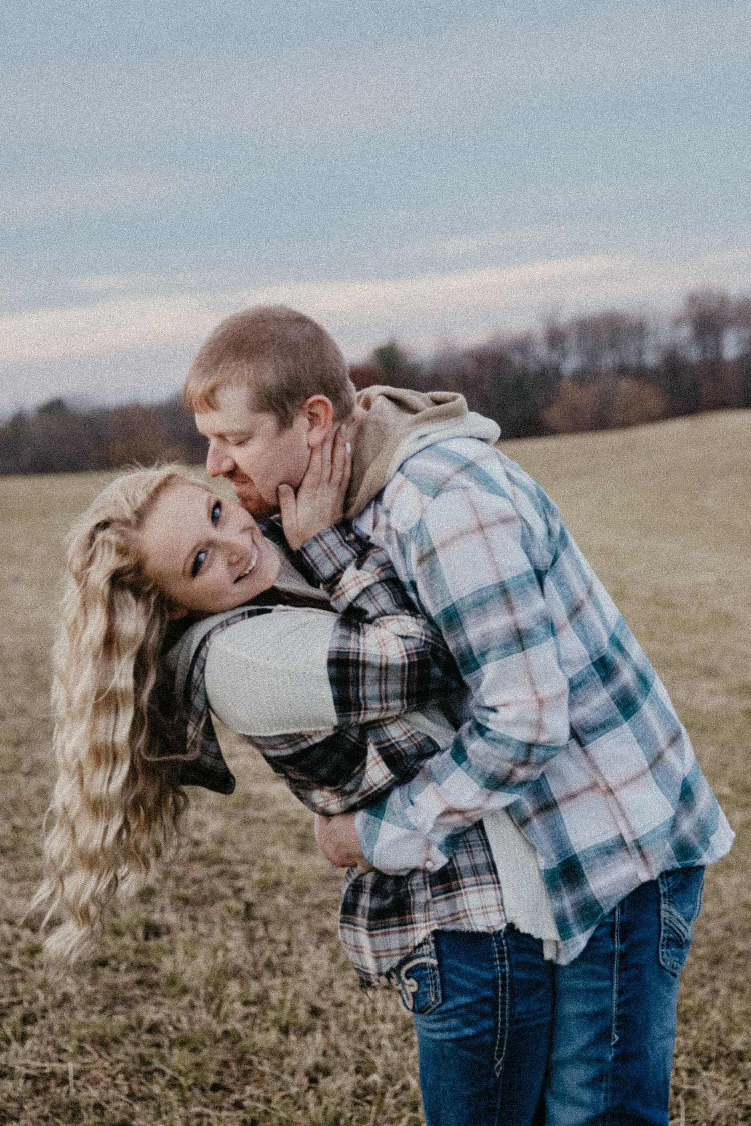 A couple shares a romantic moment in a field, man kissing woman's cheek as she smiles, both in flannel and casual wear.