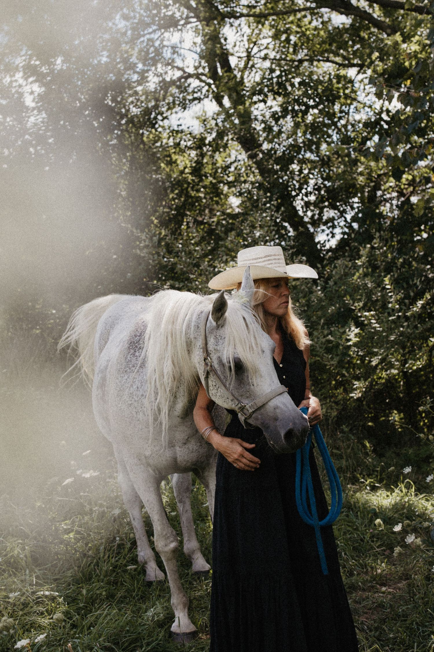 Woman in black dress and cowboy hat stands beside a white horse in a misty forest setting.