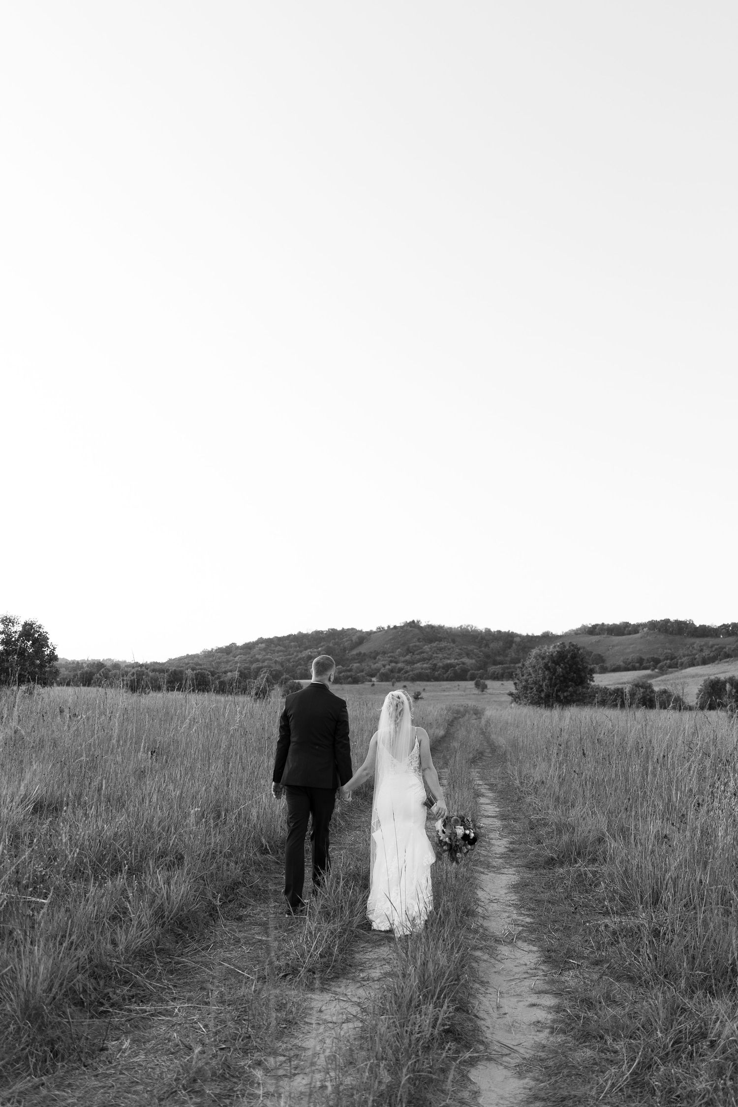 Black and white photo of a bride and groom walking hand in hand down a rural path through open fields.