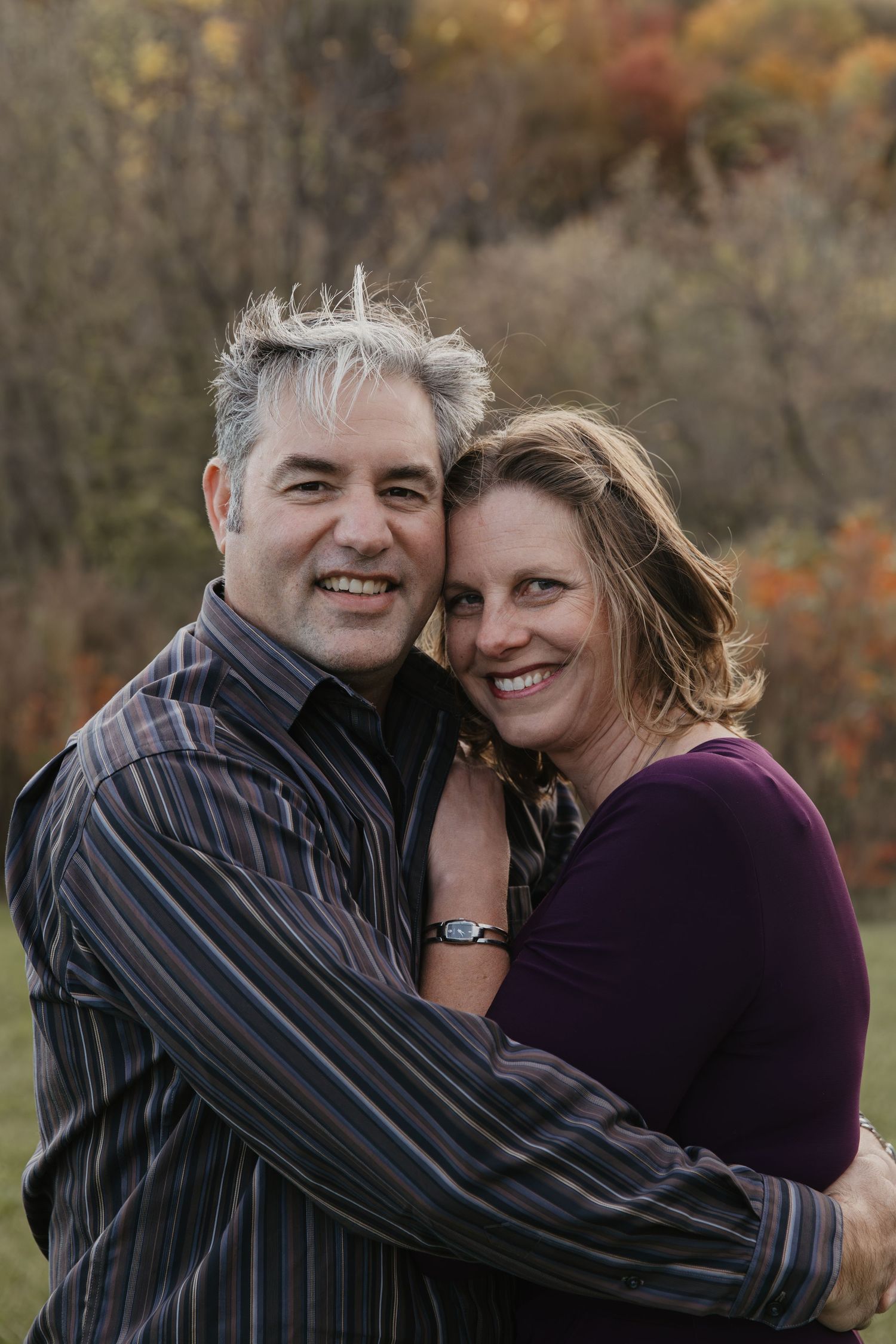 A smiling couple embraces outdoors in autumn, the man with gray hair in a striped shirt and woman in a purple top.