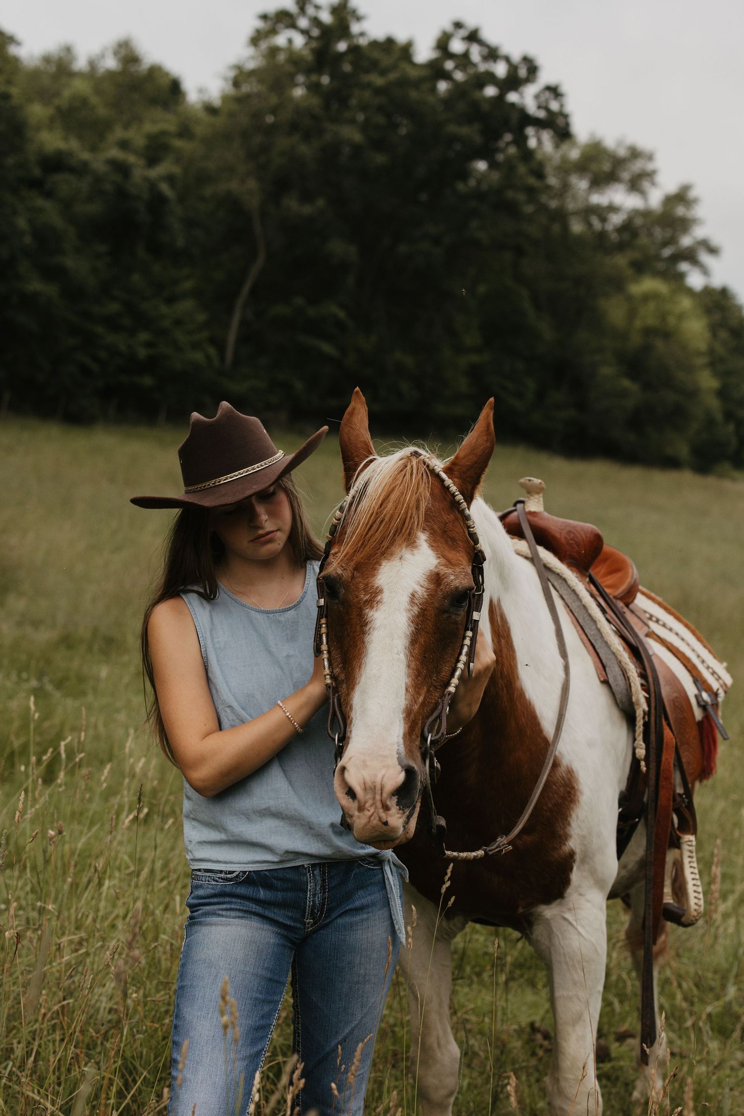 Woman in a brown cowboy hat and jeans stands beside a saddled brown and white paint horse in a grassy field with trees behind.