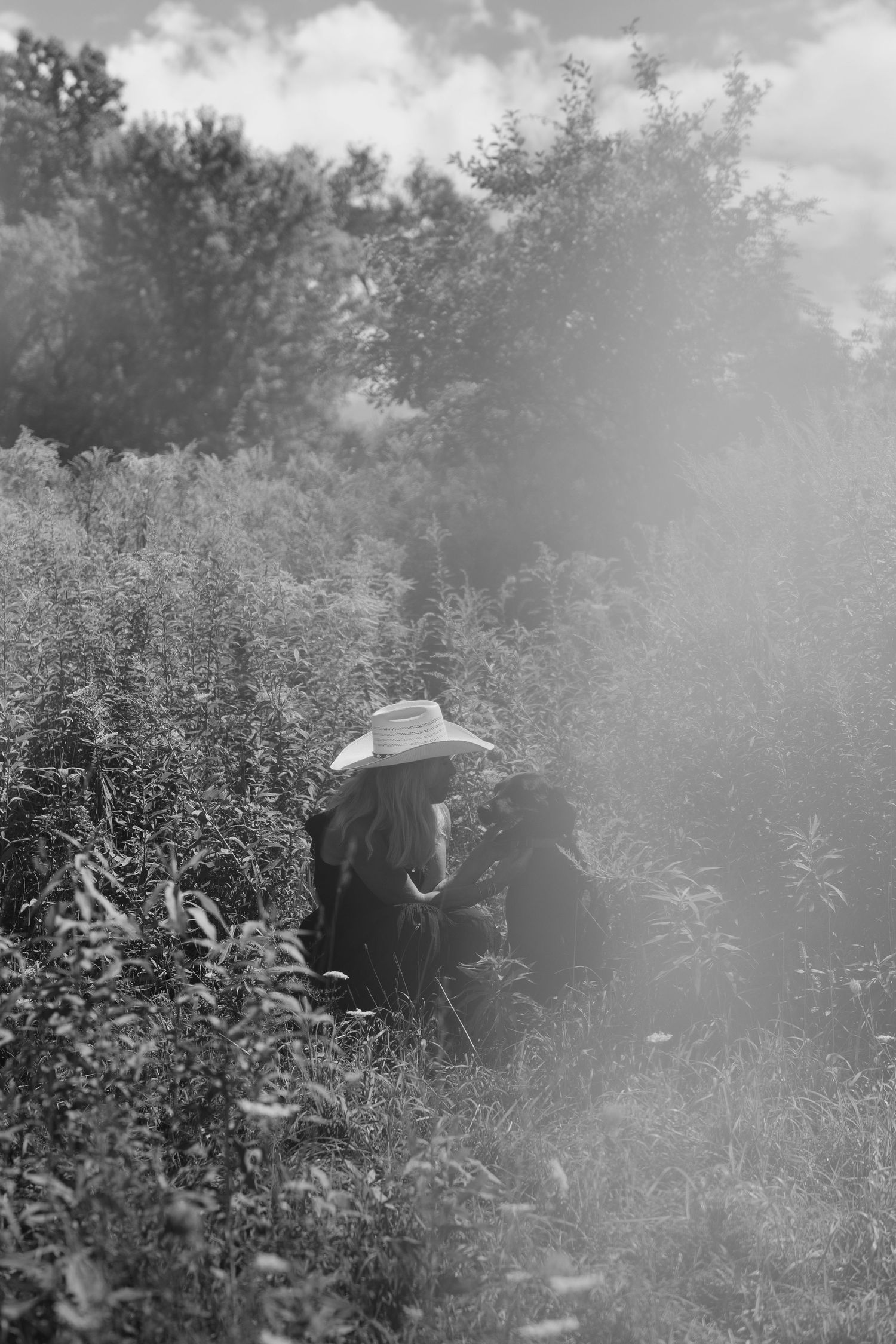 Black and white photo of a person in a wide-brim hat crouching in lush overgrown field surrounded by misty fog.