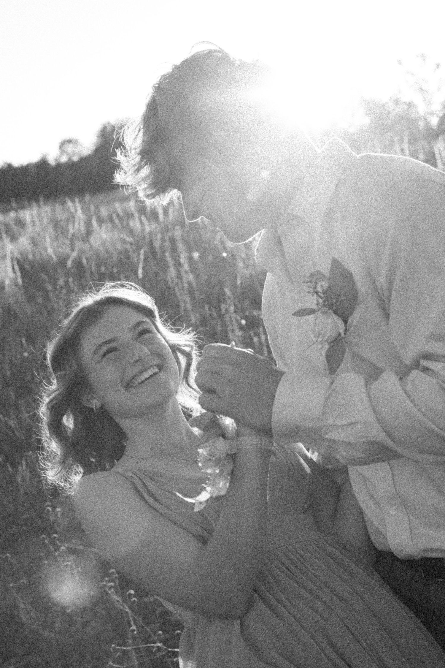 Black and white photo of a laughing couple in a sunlit field, man leaning toward smiling woman in a romantic outdoor setting.