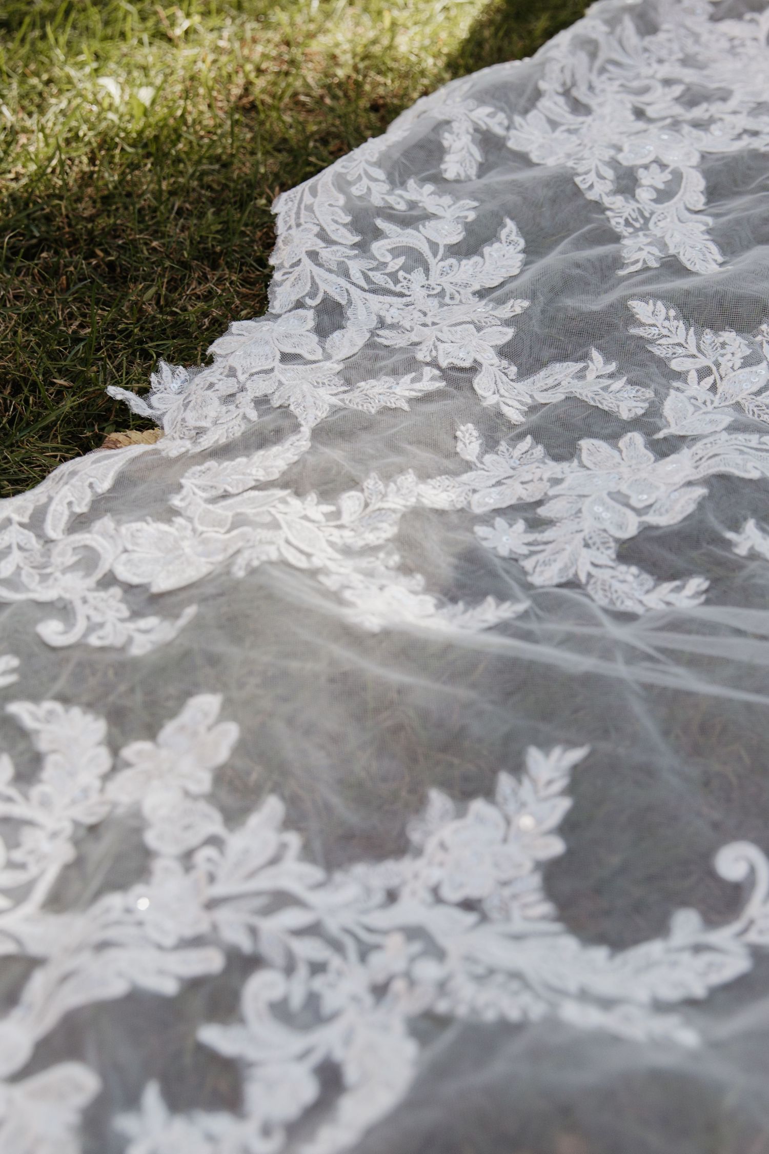 Close-up of intricate white lace bridal gown train spread on green grass outdoors.