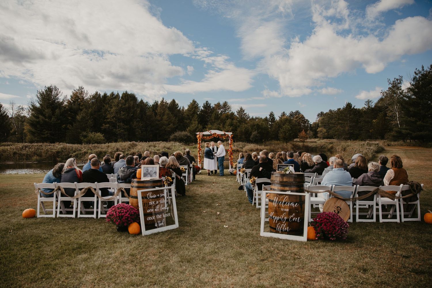 Outdoor fall wedding ceremony with guests seated in white chairs facing a wooden arch altar surrounded by autumn foliage.