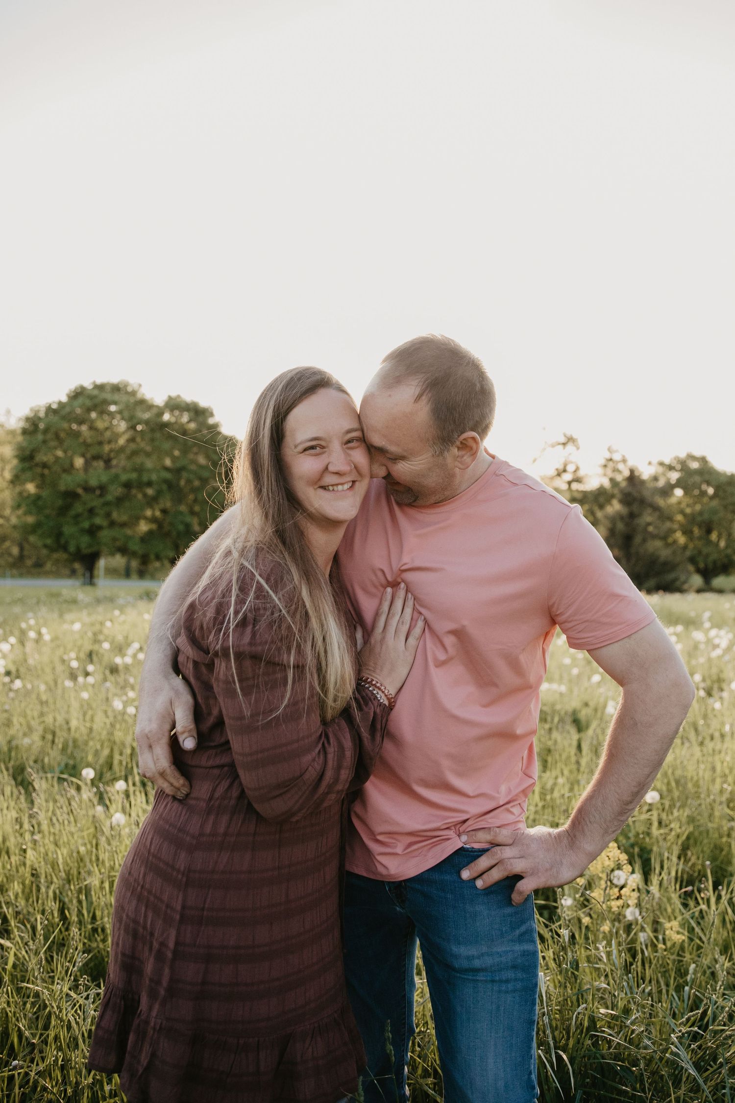 A couple embraces in a sunny meadow, man in pink shirt kissing woman in burgundy dress on the cheek.