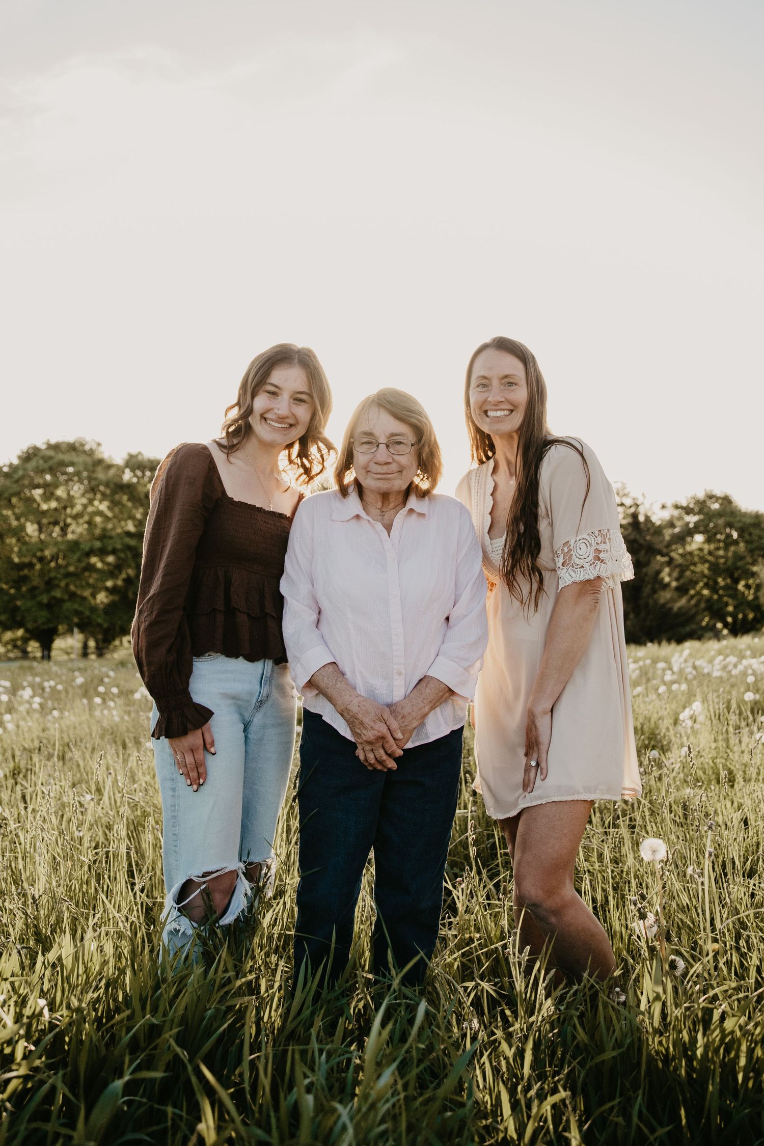 Three women of different generations smiling together in a sunlit green meadow, wearing casual summer outfits.