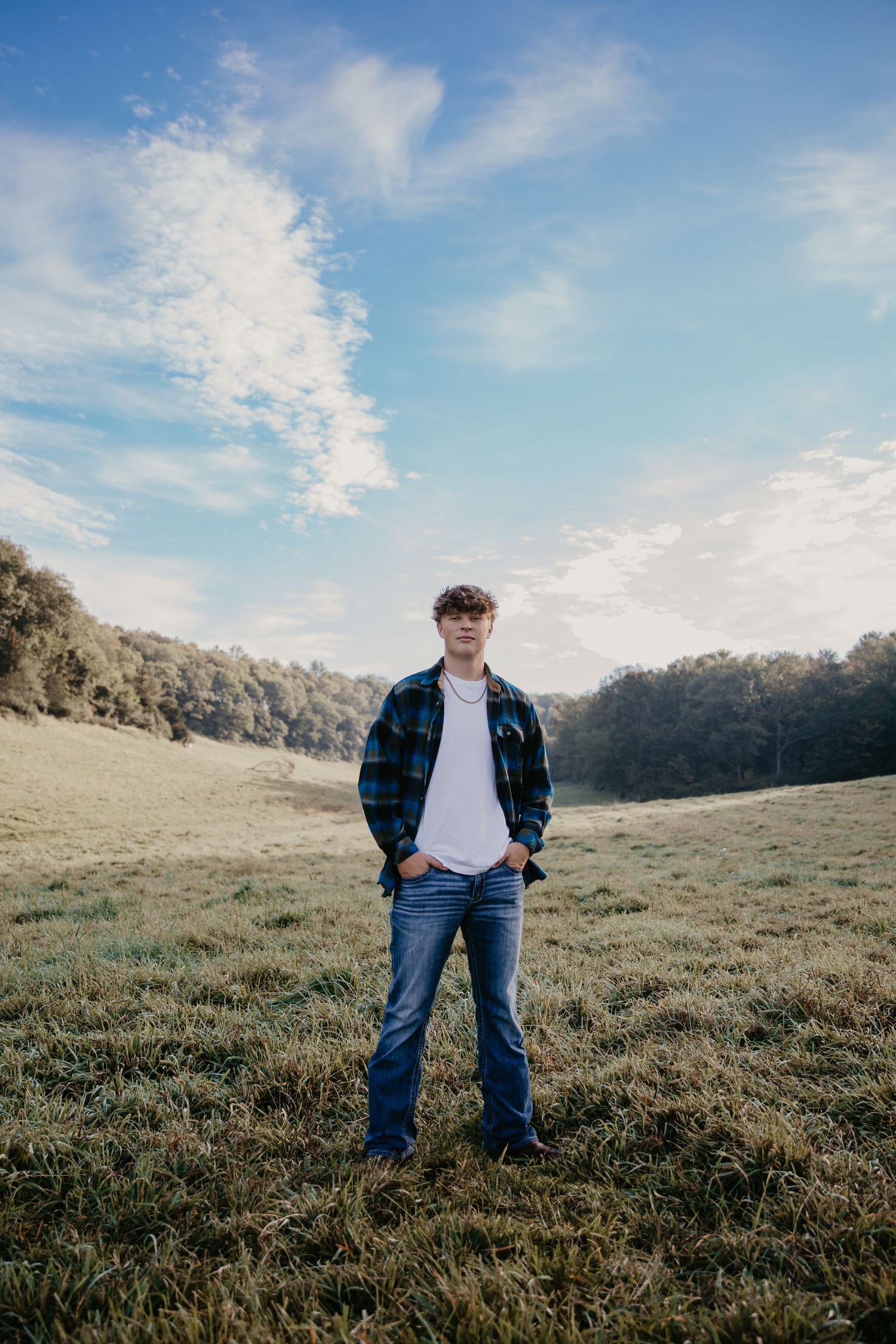 Young man in flannel shirt and jeans standing in a green meadow under a blue sky with wispy clouds.