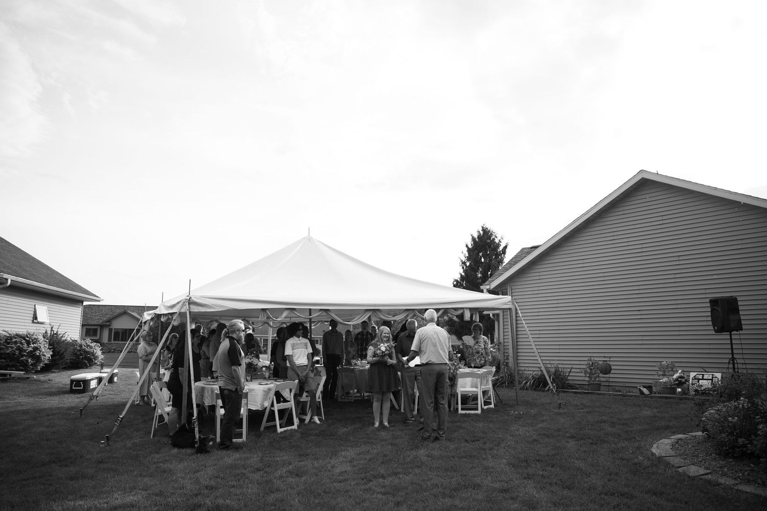 Black and white photo of guests gathered under a white tent at an outdoor backyard party or wedding reception.