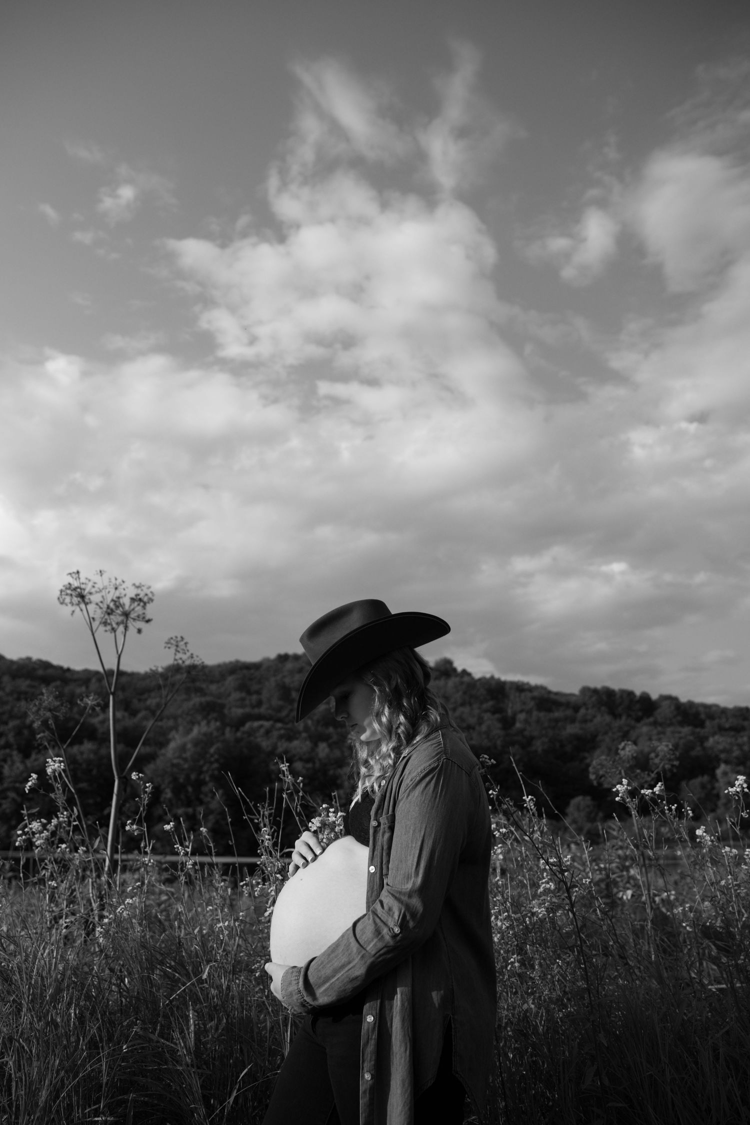 Black and white photo of pregnant woman in wide-brim hat standing in field, cradling baby bump under dramatic cloudy sky.