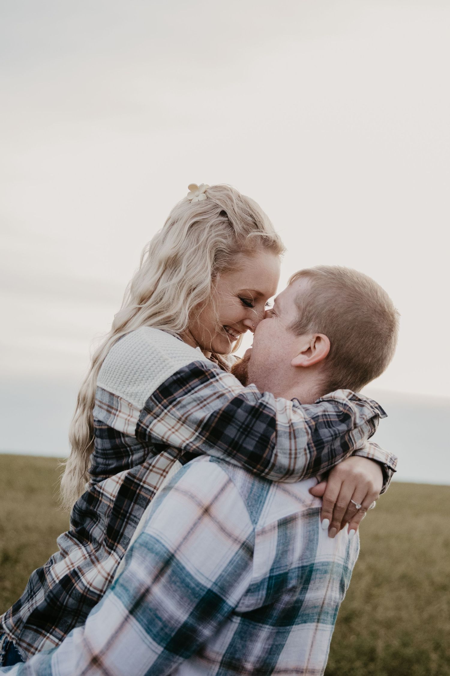 A couple in plaid shirts share a romantic forehead-to-forehead moment outdoors in a field under a cloudy sky.