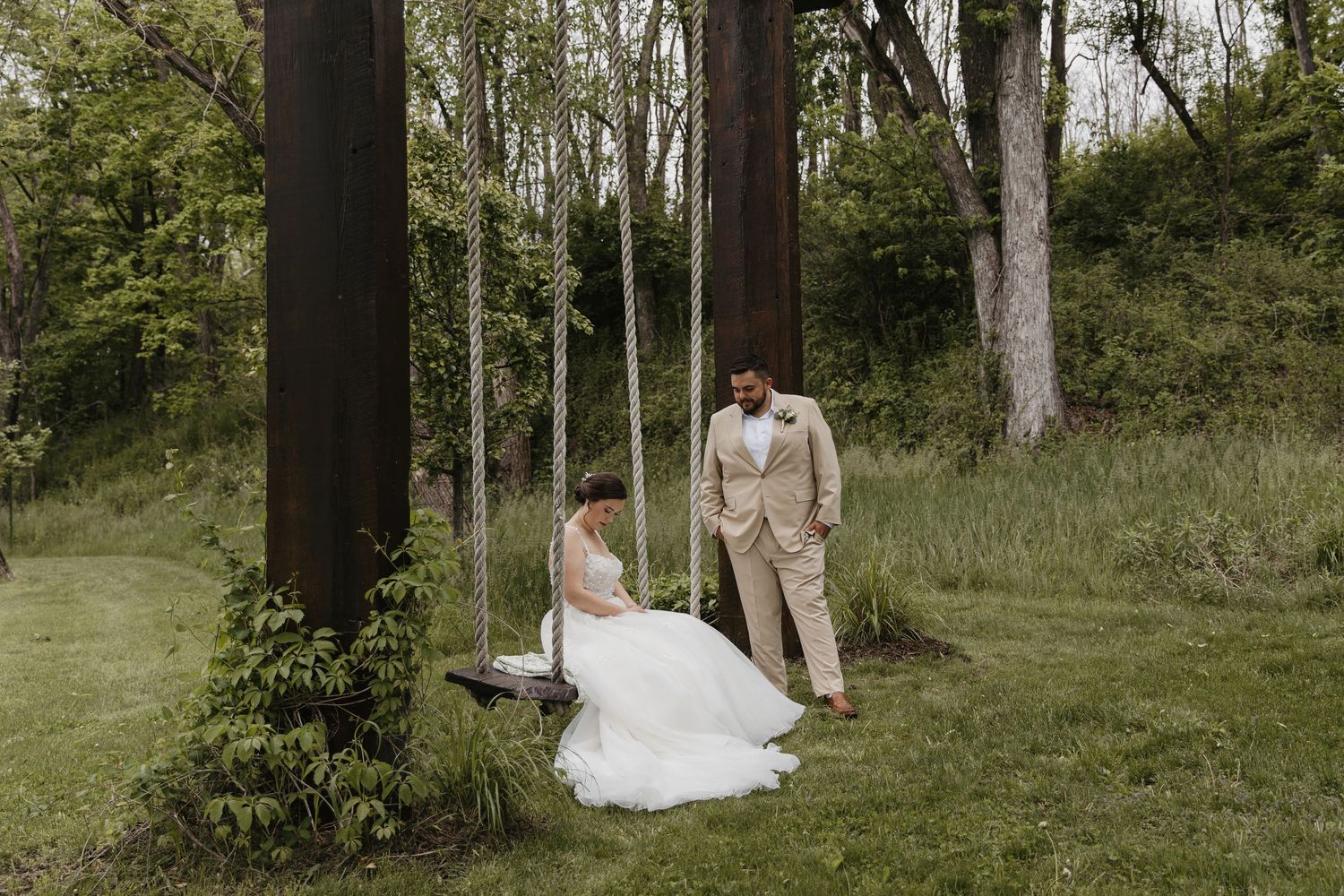 Bride in white gown sits on a wooden swing while groom in tan suit stands beside her in a lush green garden.