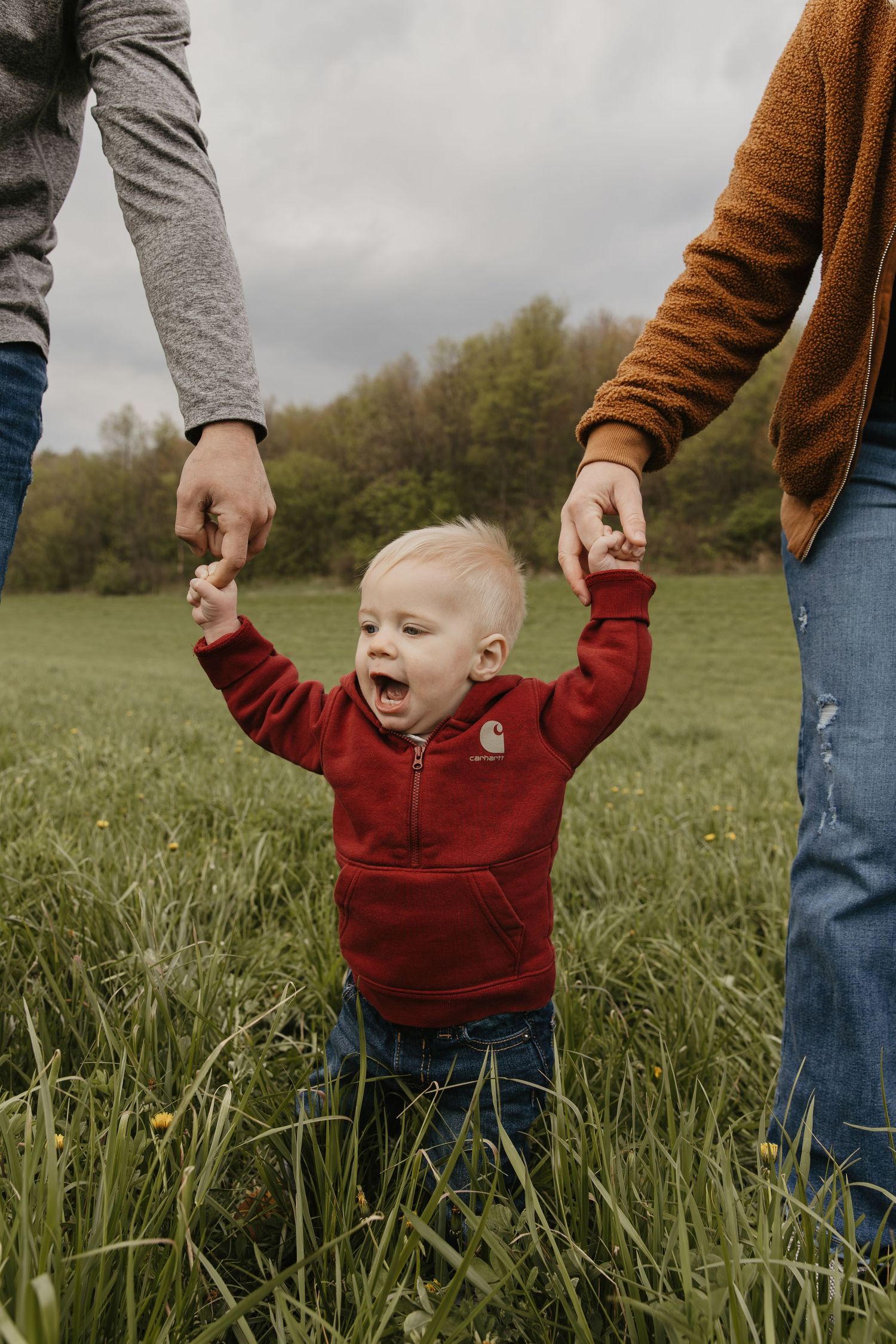 Toddler in red hoodie holds parents hands in green field, laughing outdoors on cloudy day.