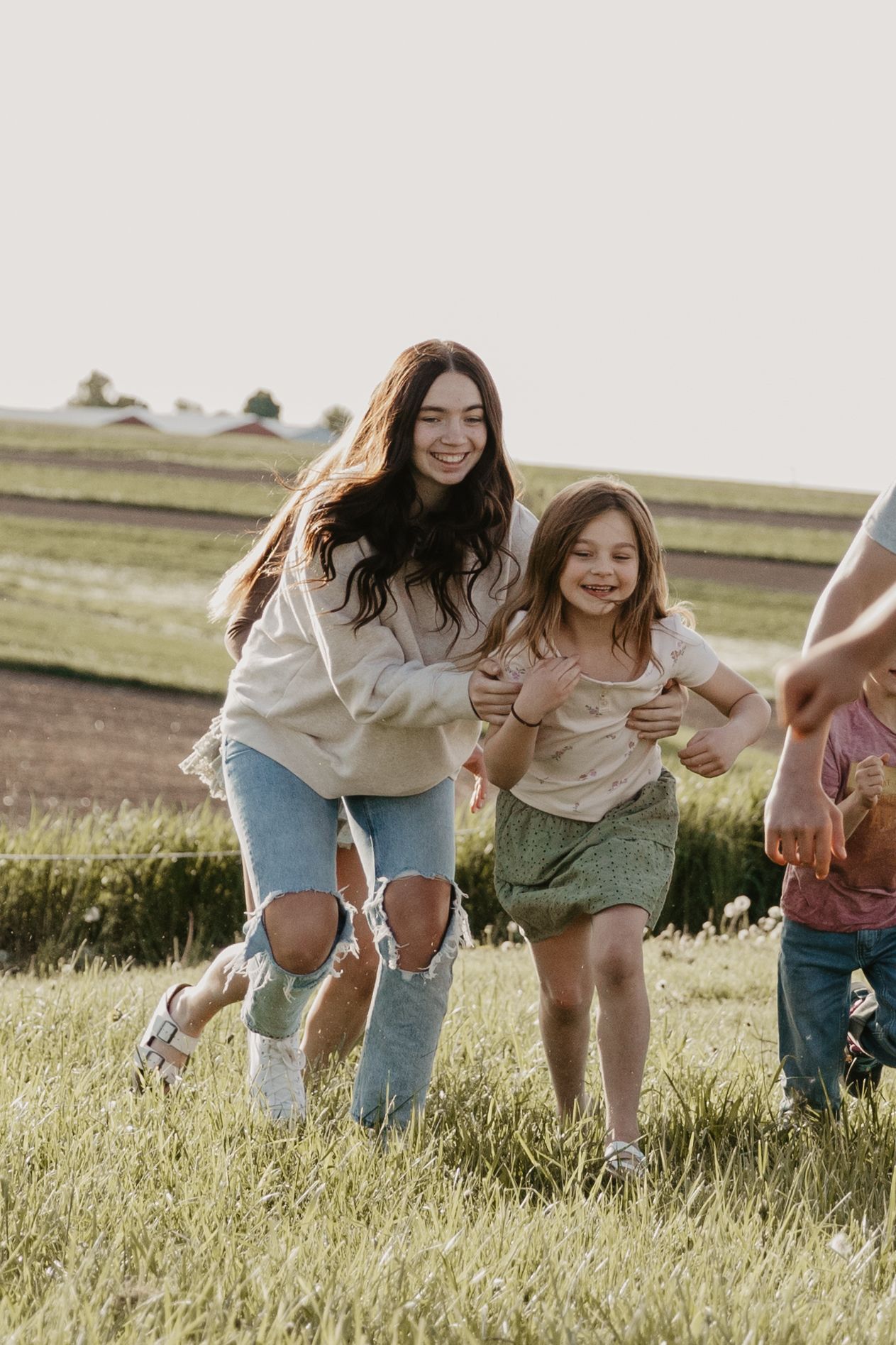 Two girls laughing and running together through a green field on a sunny day in casual outdoor clothing.