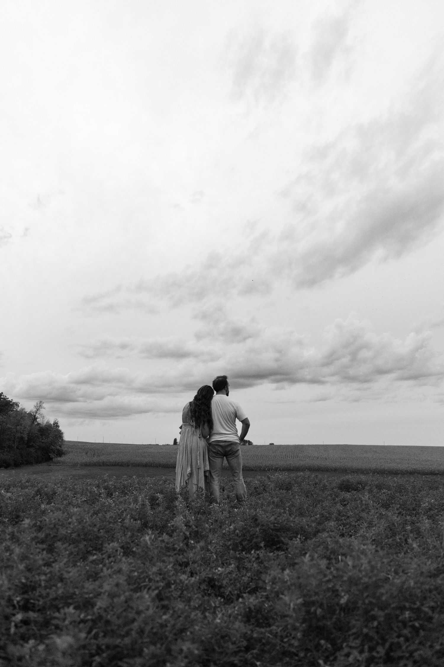 Black and white photo of a couple standing together in a field, gazing at a dramatic cloudy sky on the horizon.