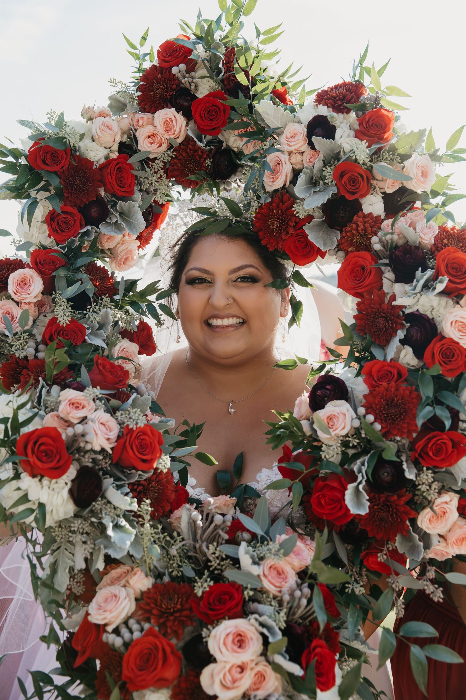 Smiling bride framed by a circular arch of red, pink, and burgundy roses with eucalyptus greenery at her wedding.