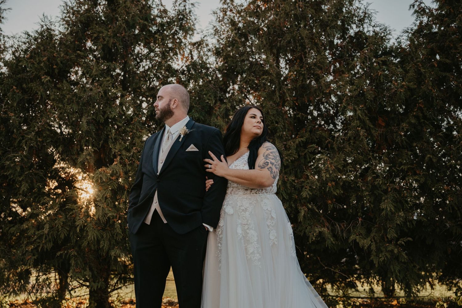 Bride in lace gown and groom in dark suit pose romantically before lush green trees at golden hour wedding.