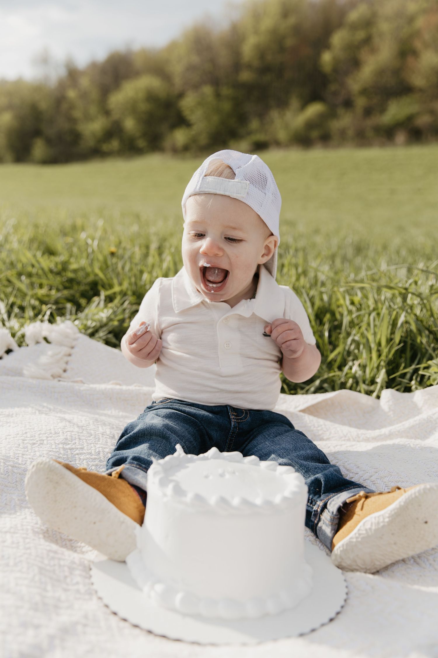 Excited baby boy in white cap and jeans sits on blanket outdoors with white smash cake for first birthday.