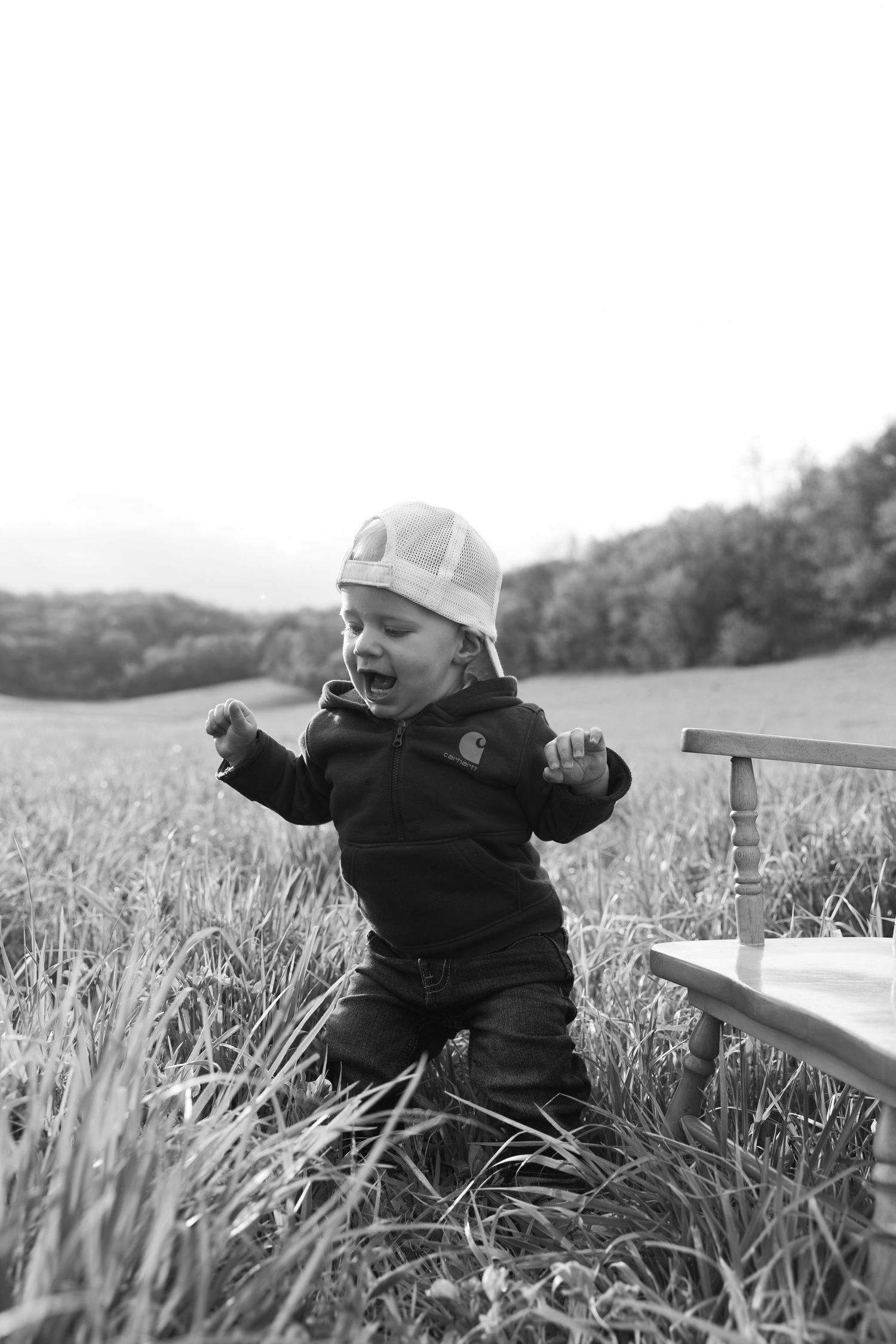 Black and white photo of a toddler in a cap and jacket playing in tall grass in a rural field.